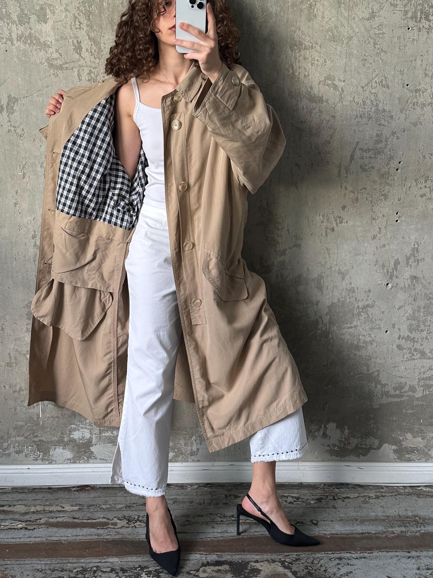 A woman in a beige trench coat and white pants is taking a selfie with a white phone, standing against a concrete wall.