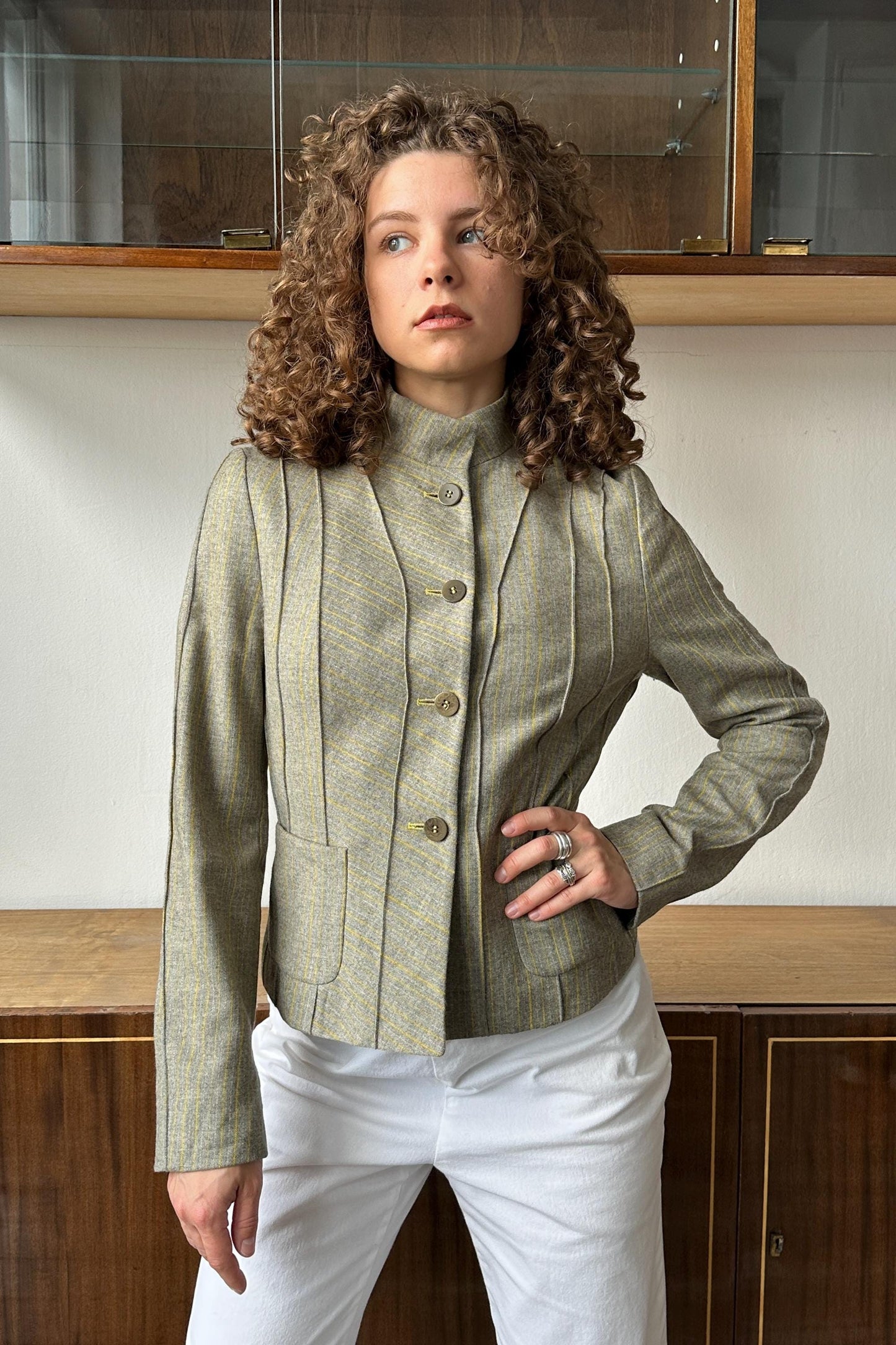 A woman with curly hair wearing a gray jacket and white pants stands in front of a wooden cabinet.