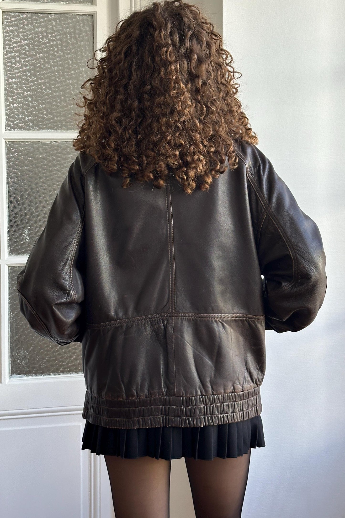 a person with curly hair wearing a brown leather jacket and a black skirt, standing in front of a window.