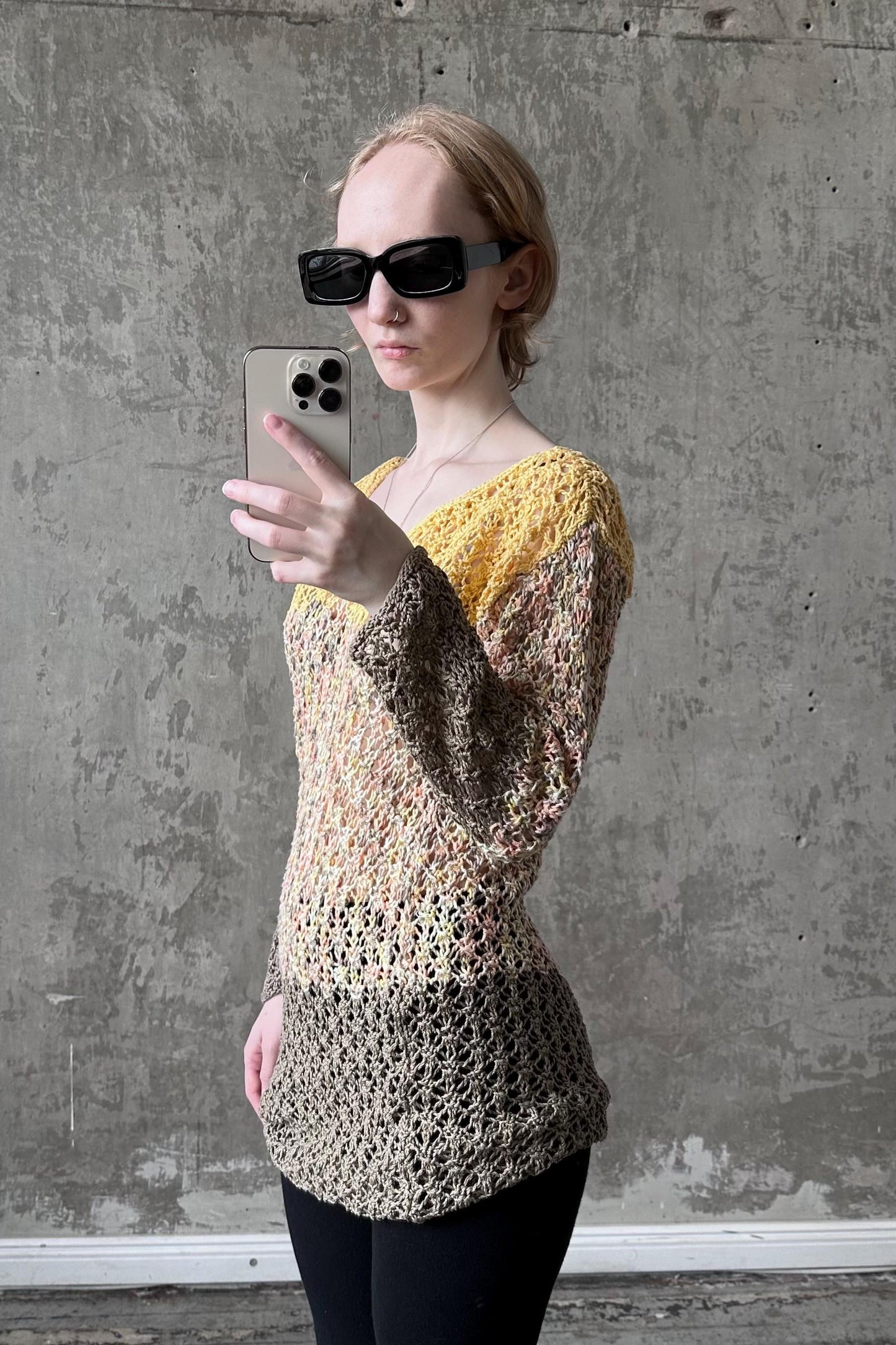 A woman wearing a yellow and gray sweater and sunglasses is taking a selfie with a smartphone against a concrete wall.