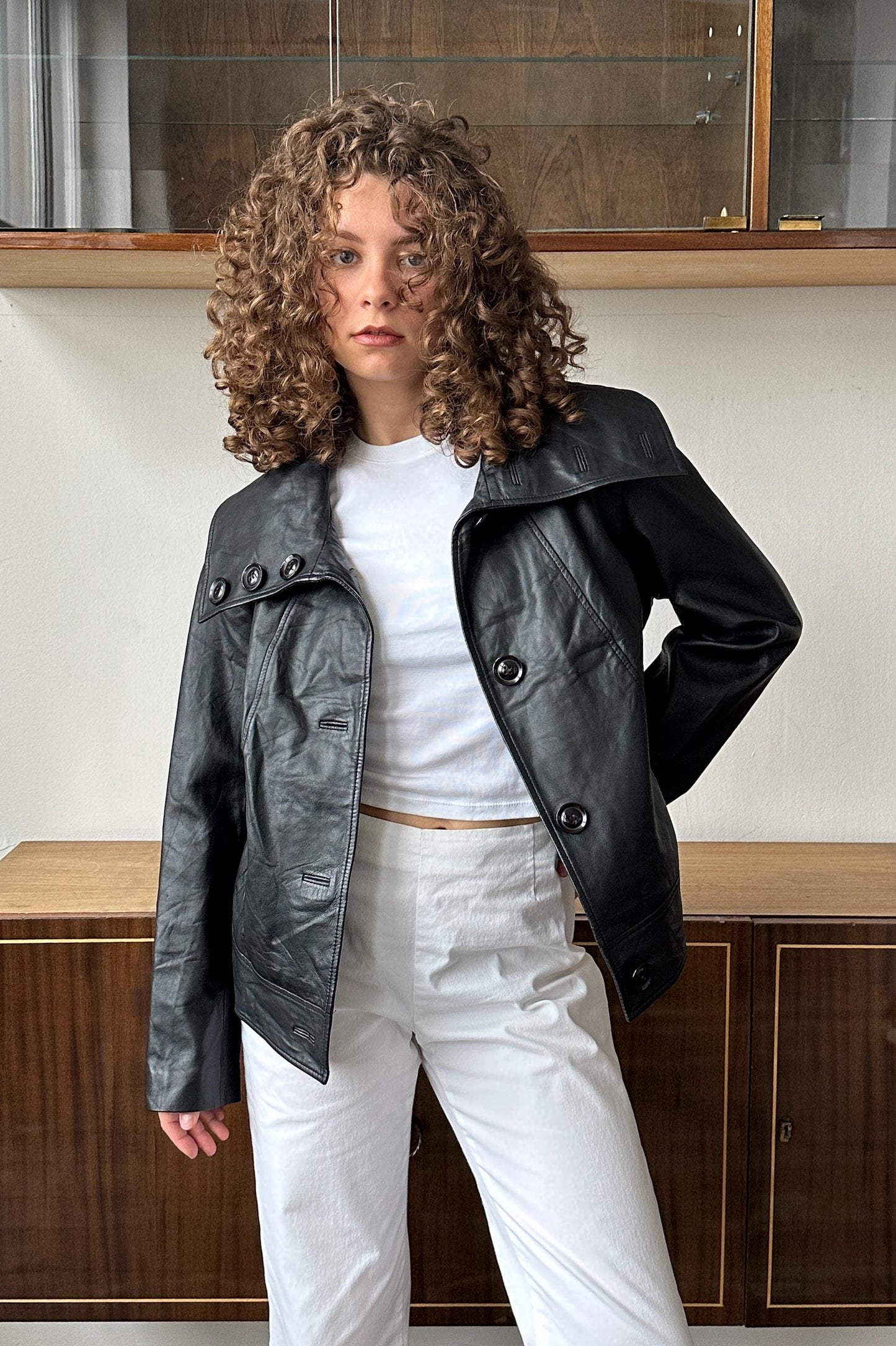 A woman with curly hair wearing a black leather jacket and white pants stands in front of a wooden cabinet.