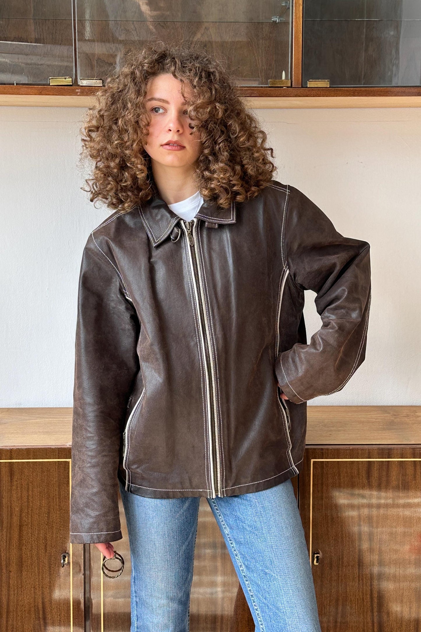 A young woman with curly hair wearing a brown leather jacket stands in front of a wooden cabinet.