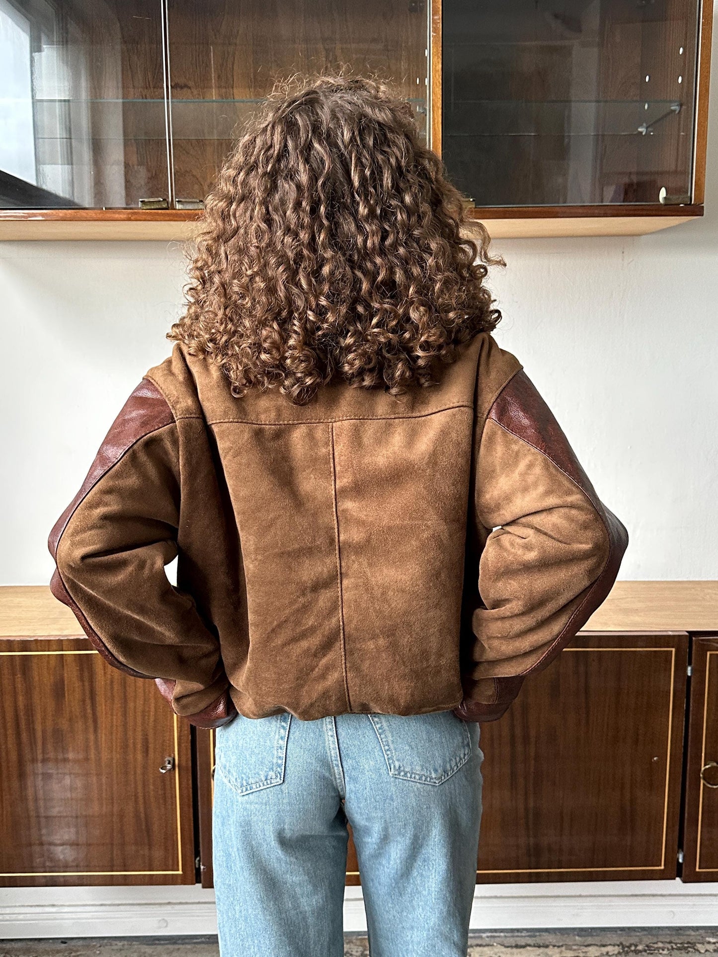 a person with curly hair wearing a brown jacket and blue jeans, standing in front of a wooden cabinet.