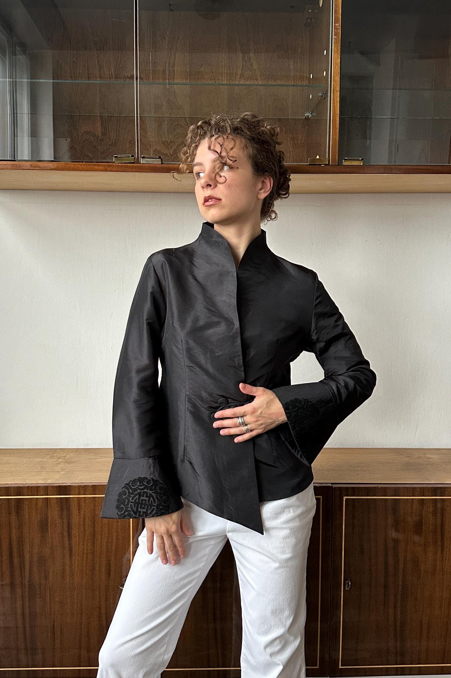A woman wearing a black jacket with a high collar and long sleeves stands in front of a wooden cabinet.