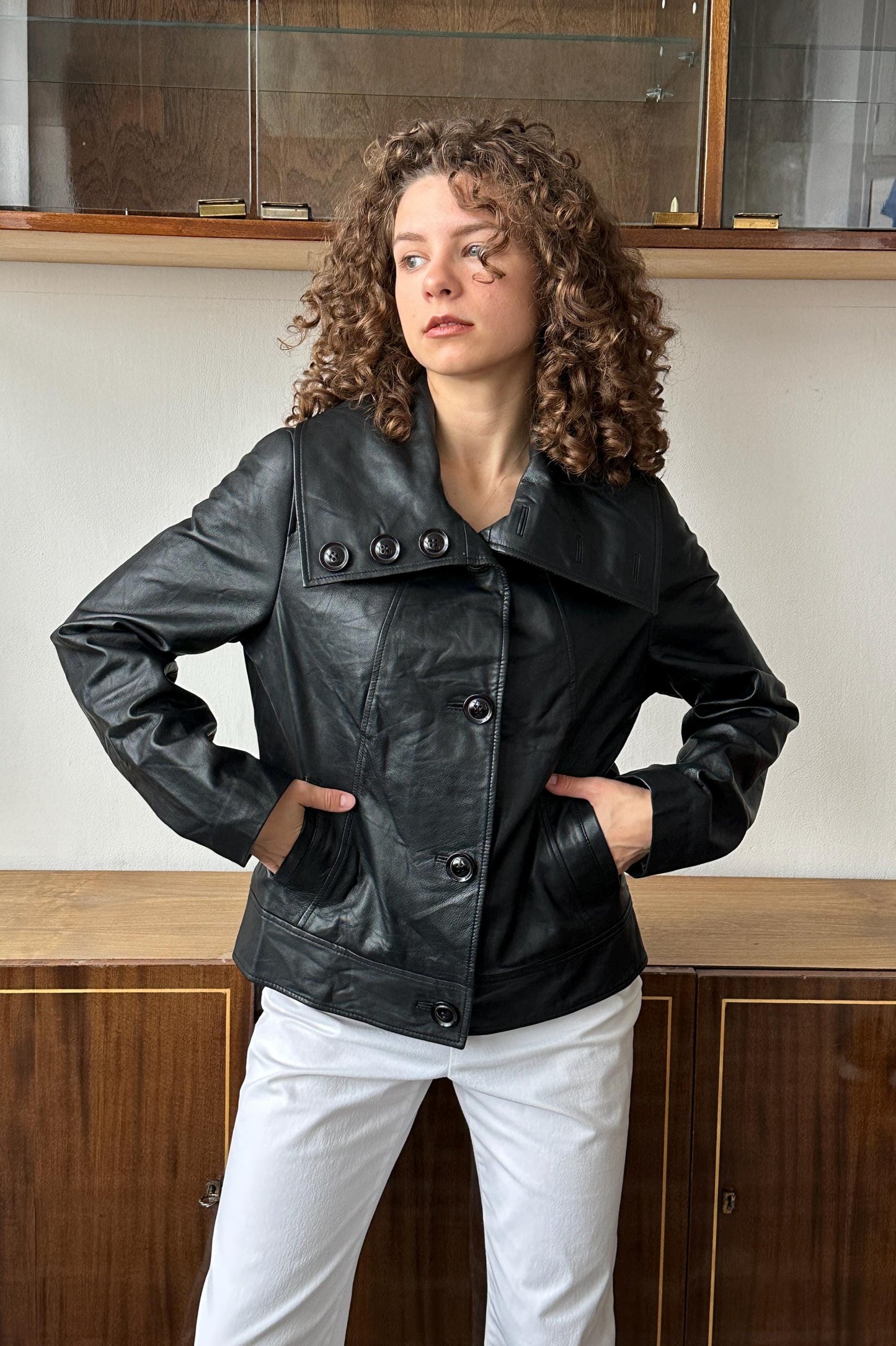A woman with curly hair wearing a black leather jacket stands in front of a wooden cabinet.