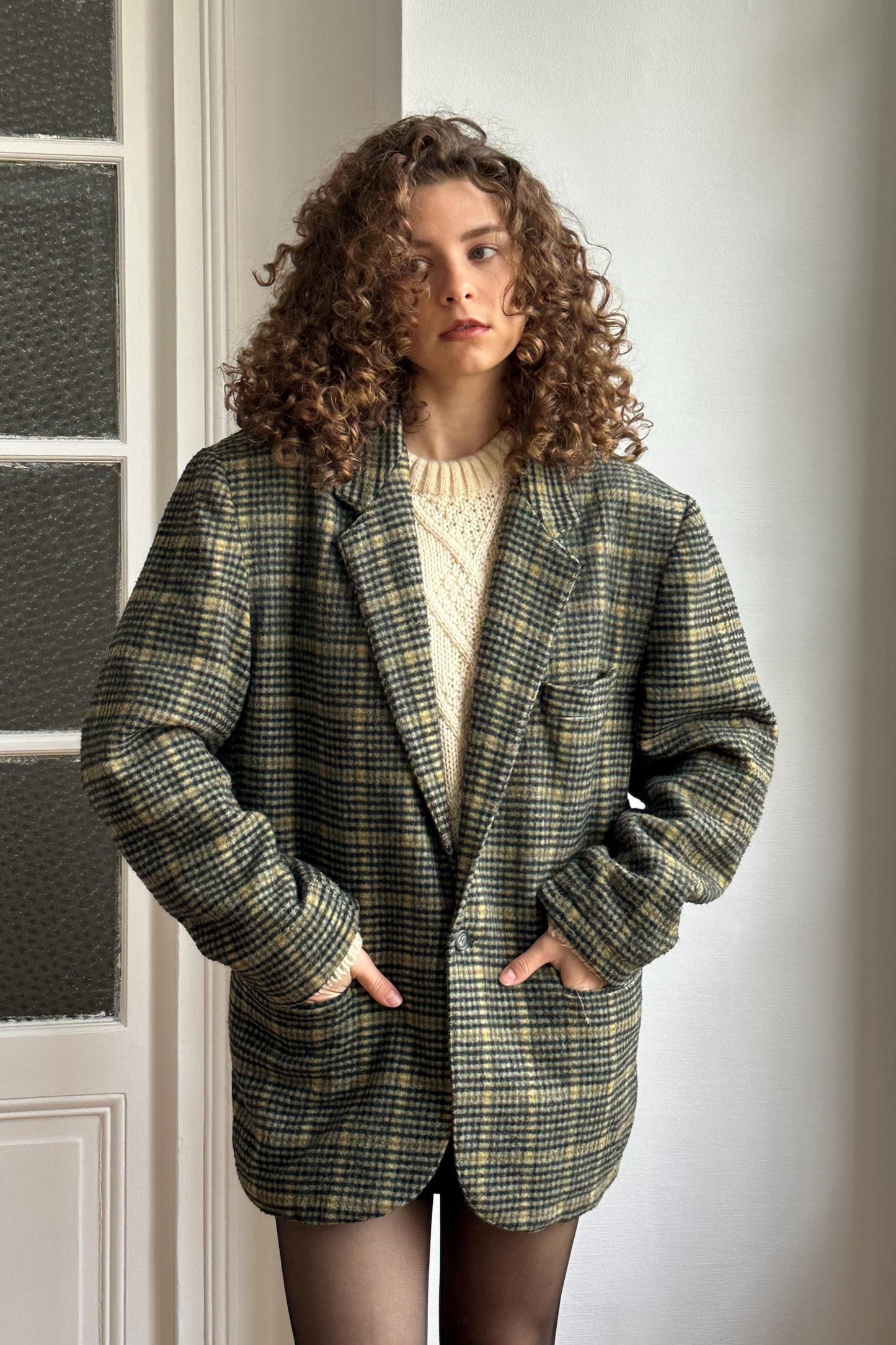A woman with curly hair wearing a plaid jacket stands in front of a white door.