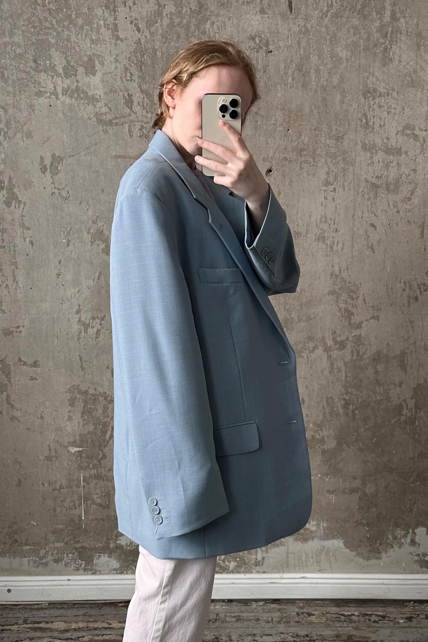 A person is taking a selfie with a smartphone, wearing a light blue blazer against a textured concrete wall background.