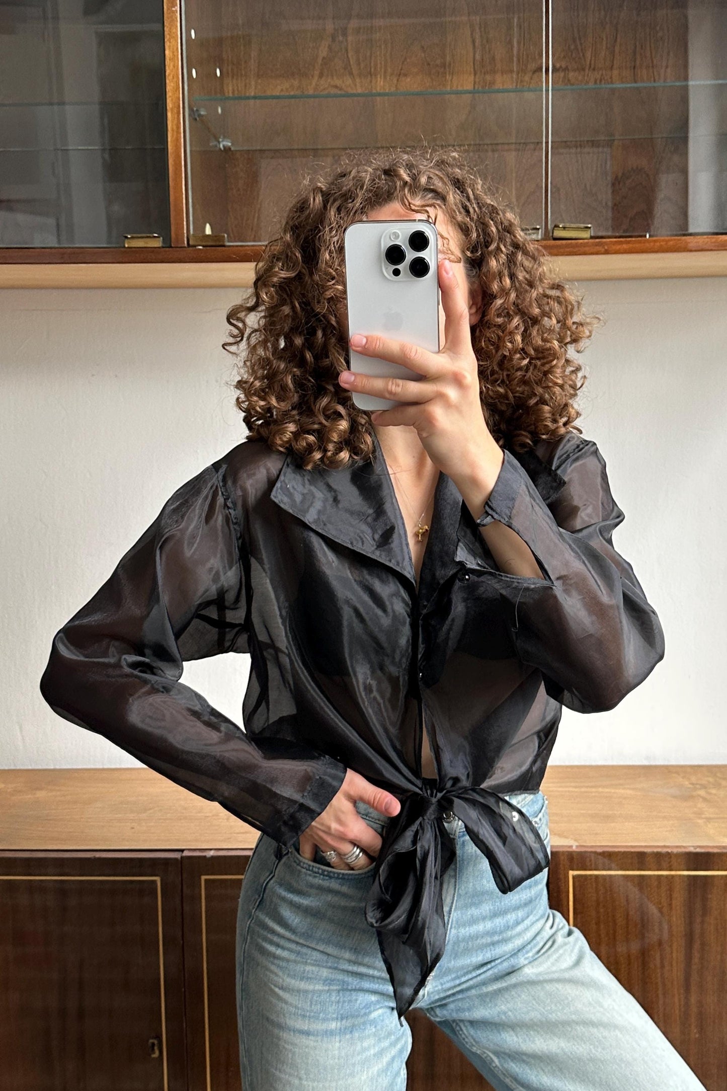 A person with curly hair is taking a selfie with a smartphone, wearing a black blouse and blue jeans.