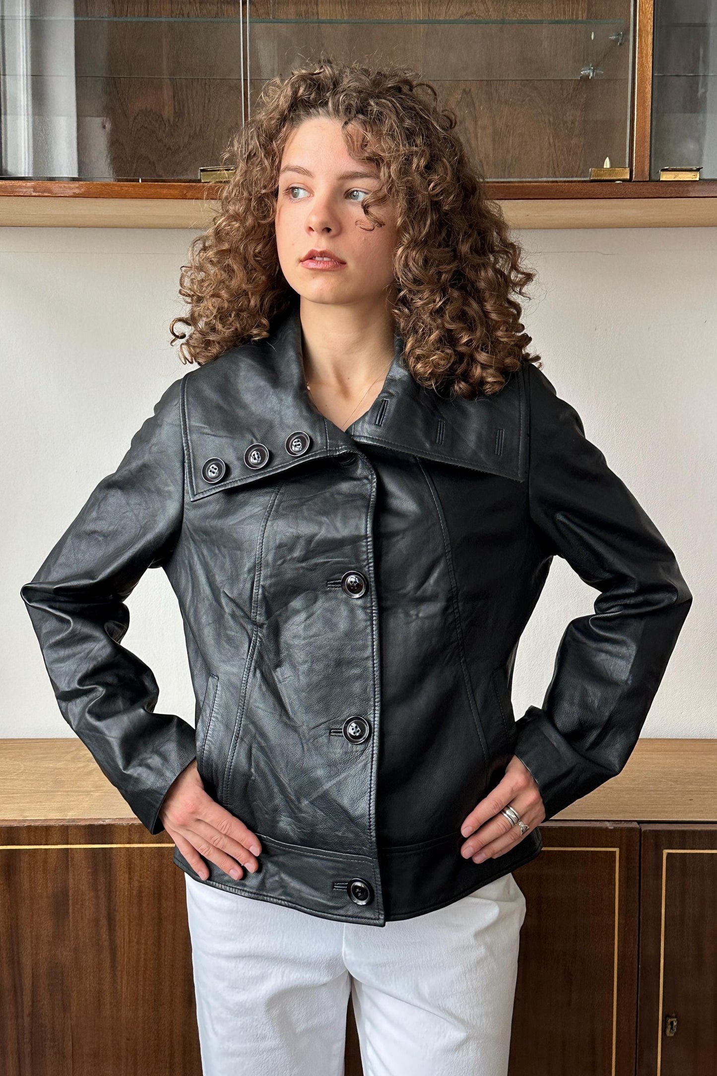 A woman with curly hair wearing a black leather jacket stands in front of a wooden cabinet.