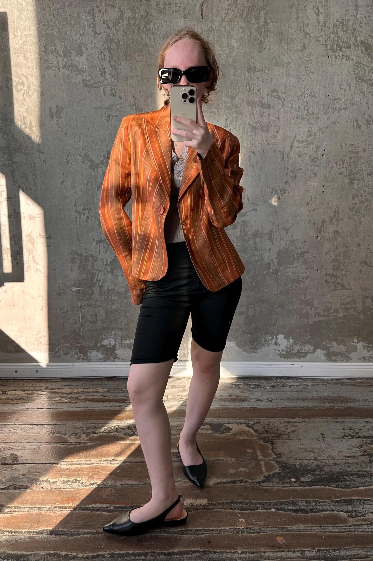 A person is taking a selfie with a smartphone, wearing a striped orange blazer, black shorts, and black shoes, standing in front of a concrete wall.
