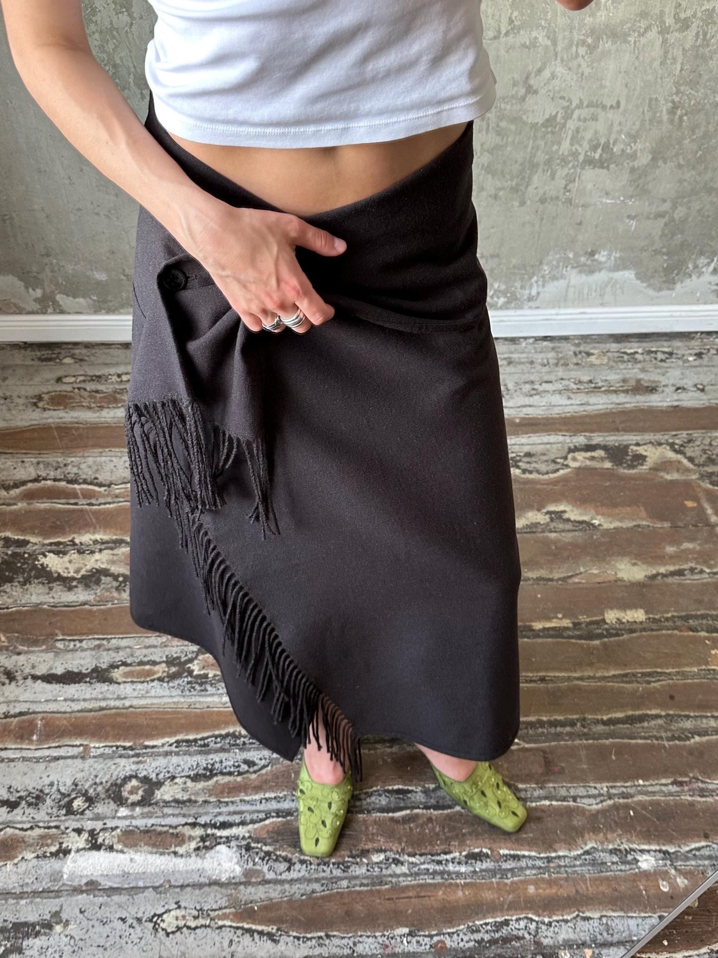 A person wearing a black skirt with fringe details and green shoes stands on a wooden floor against a concrete wall.