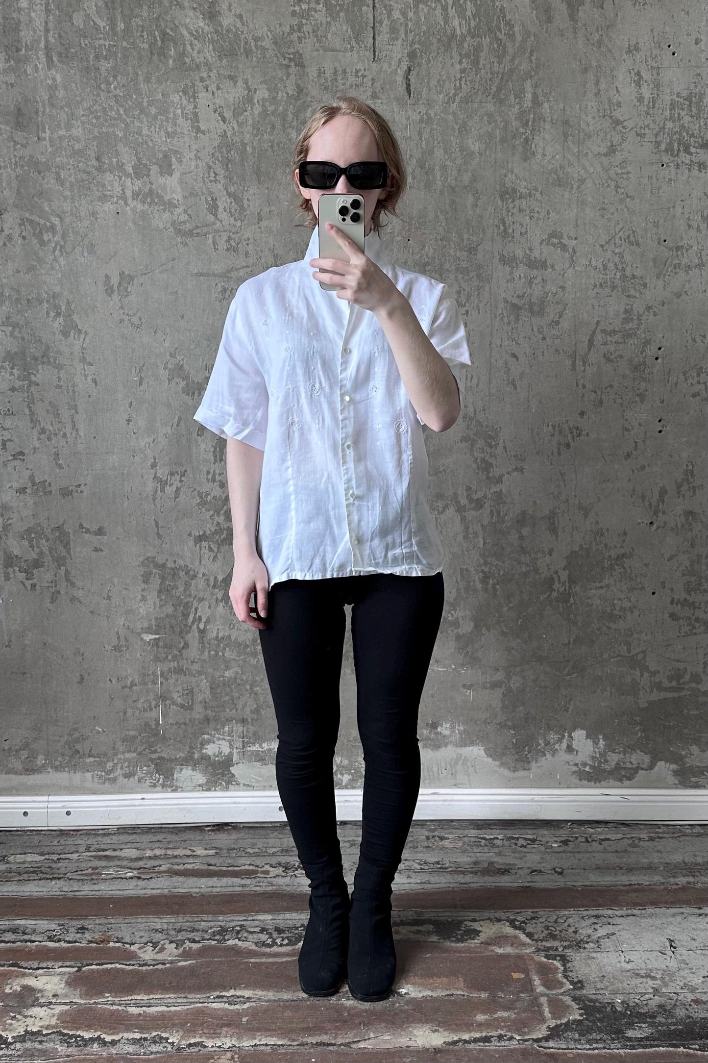 A person is taking a selfie with a white shirt, black pants, and sunglasses, standing in front of a concrete wall.