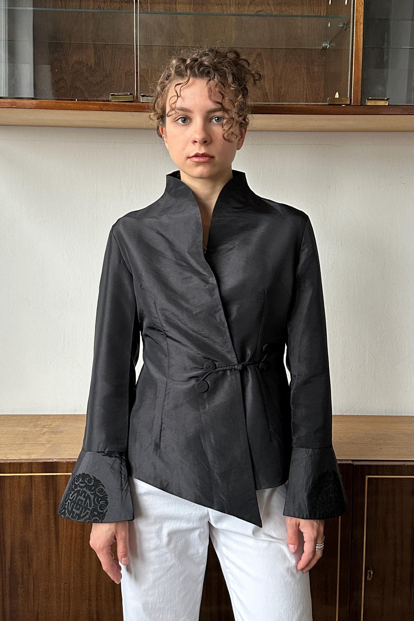 A woman wearing a black blazer with a high collar and long sleeves stands in front of a wooden cabinet.