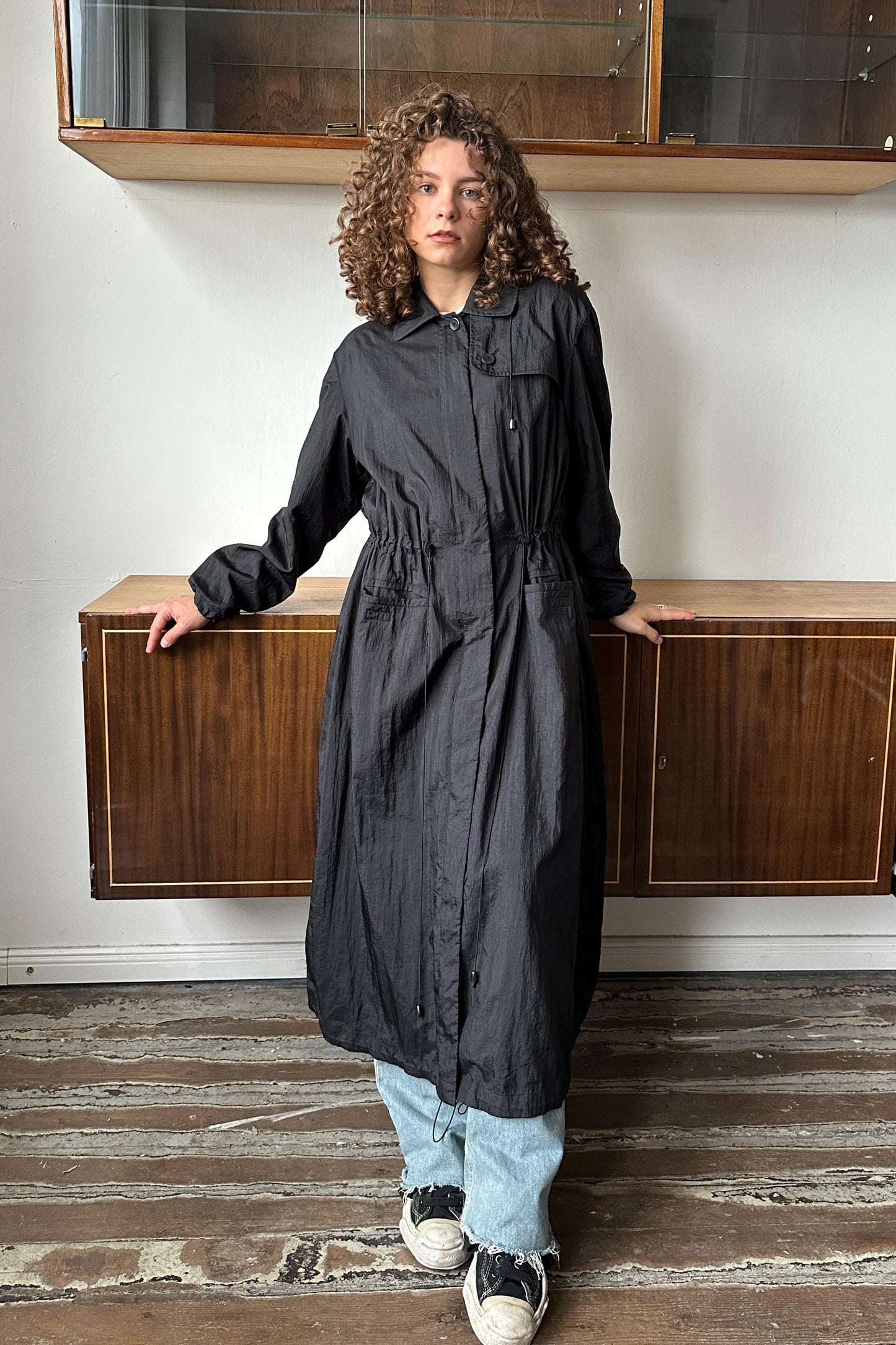 A woman with curly hair wearing a long black coat stands in a room with wooden cabinets and a wooden floor.