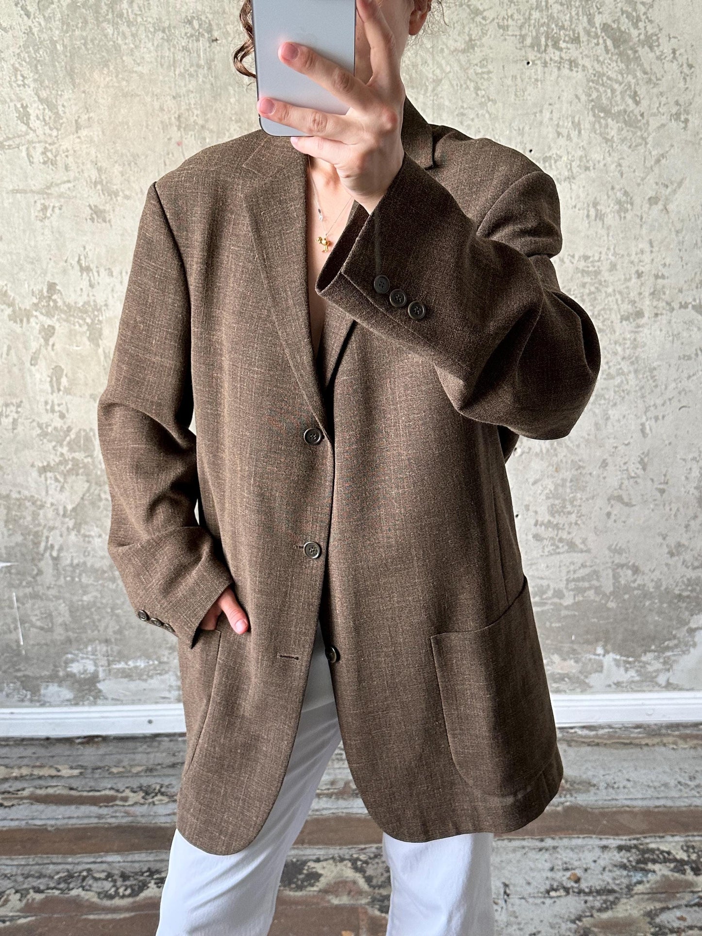 A person is taking a selfie with a brown blazer and white pants, holding a phone up to their face.