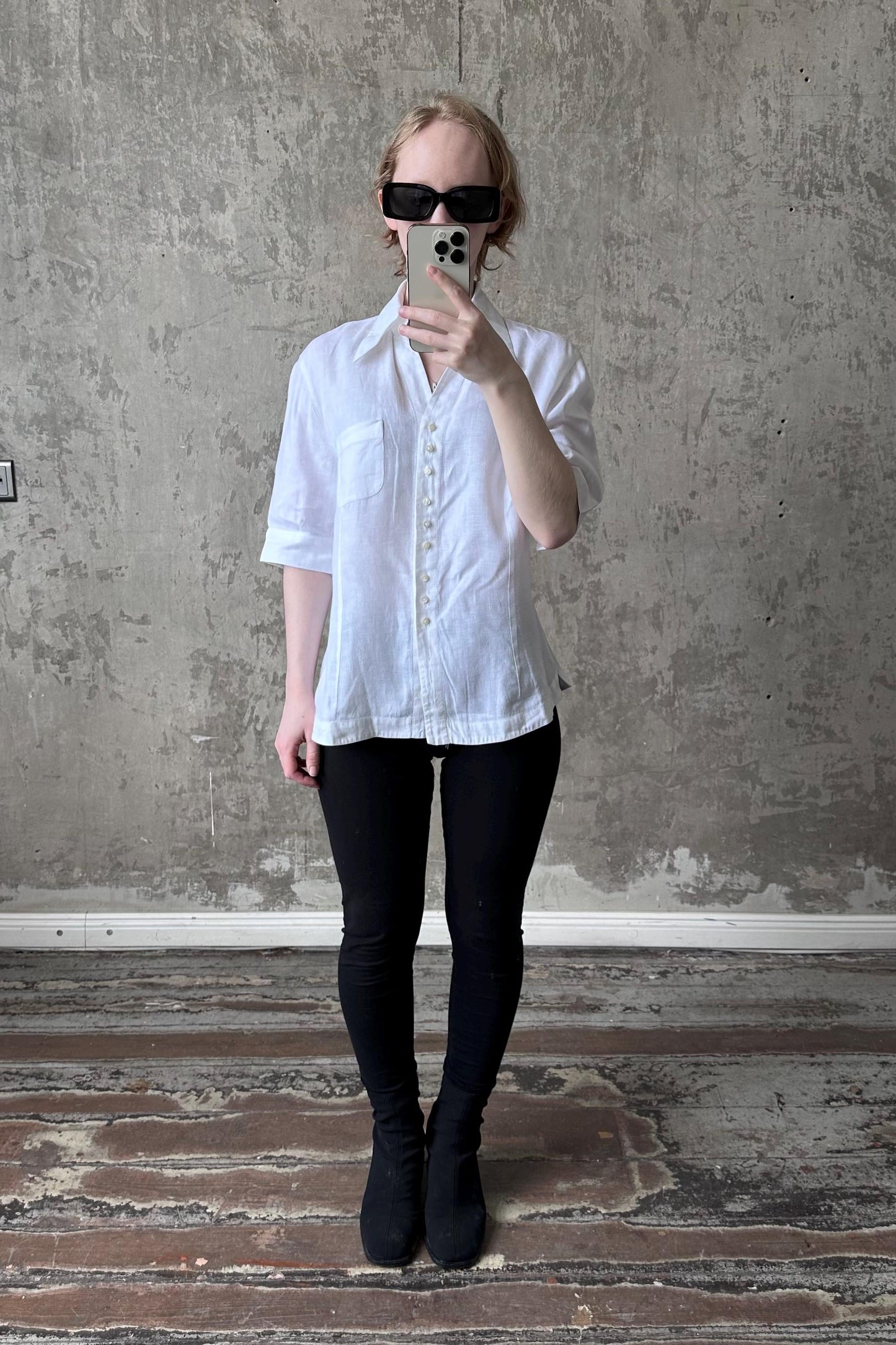 A person is taking a selfie with a white shirt, black pants, and black boots, standing in front of a concrete wall.