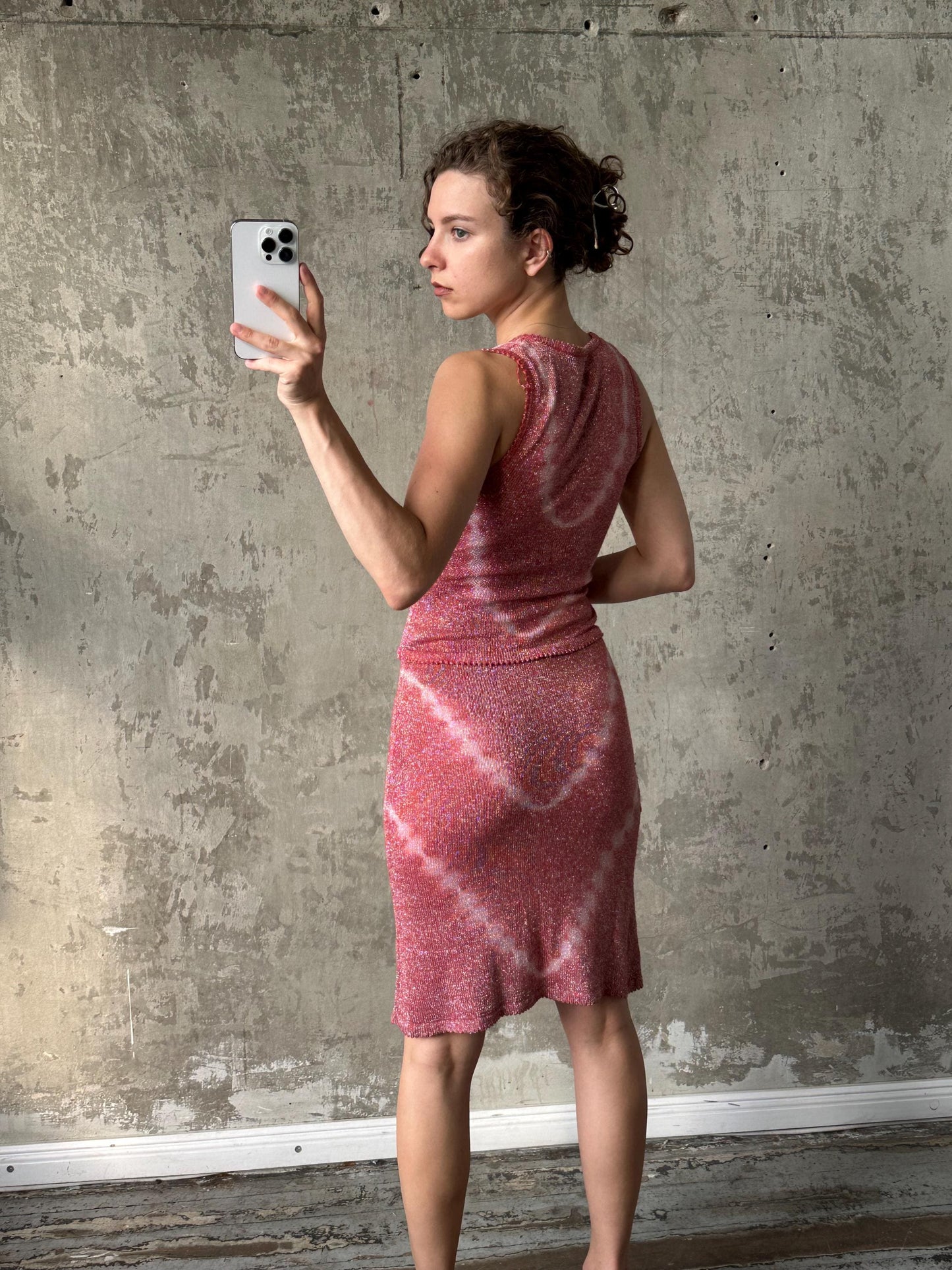 A woman in a pink dress is taking a selfie with a smartphone, standing in front of a concrete wall.