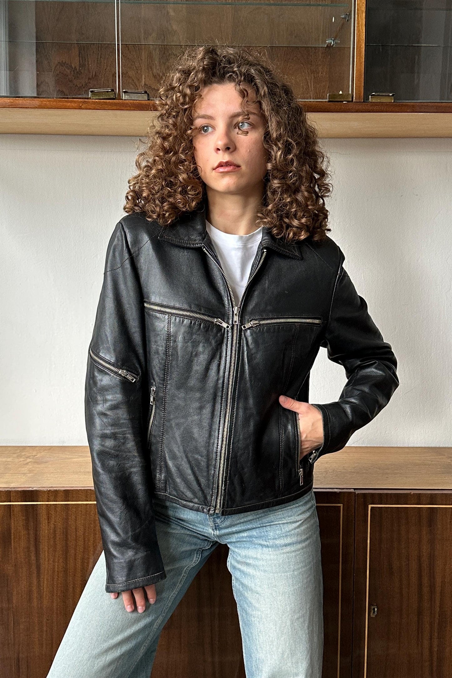 A young woman with curly hair wearing a black leather jacket stands in front of a wooden cabinet.