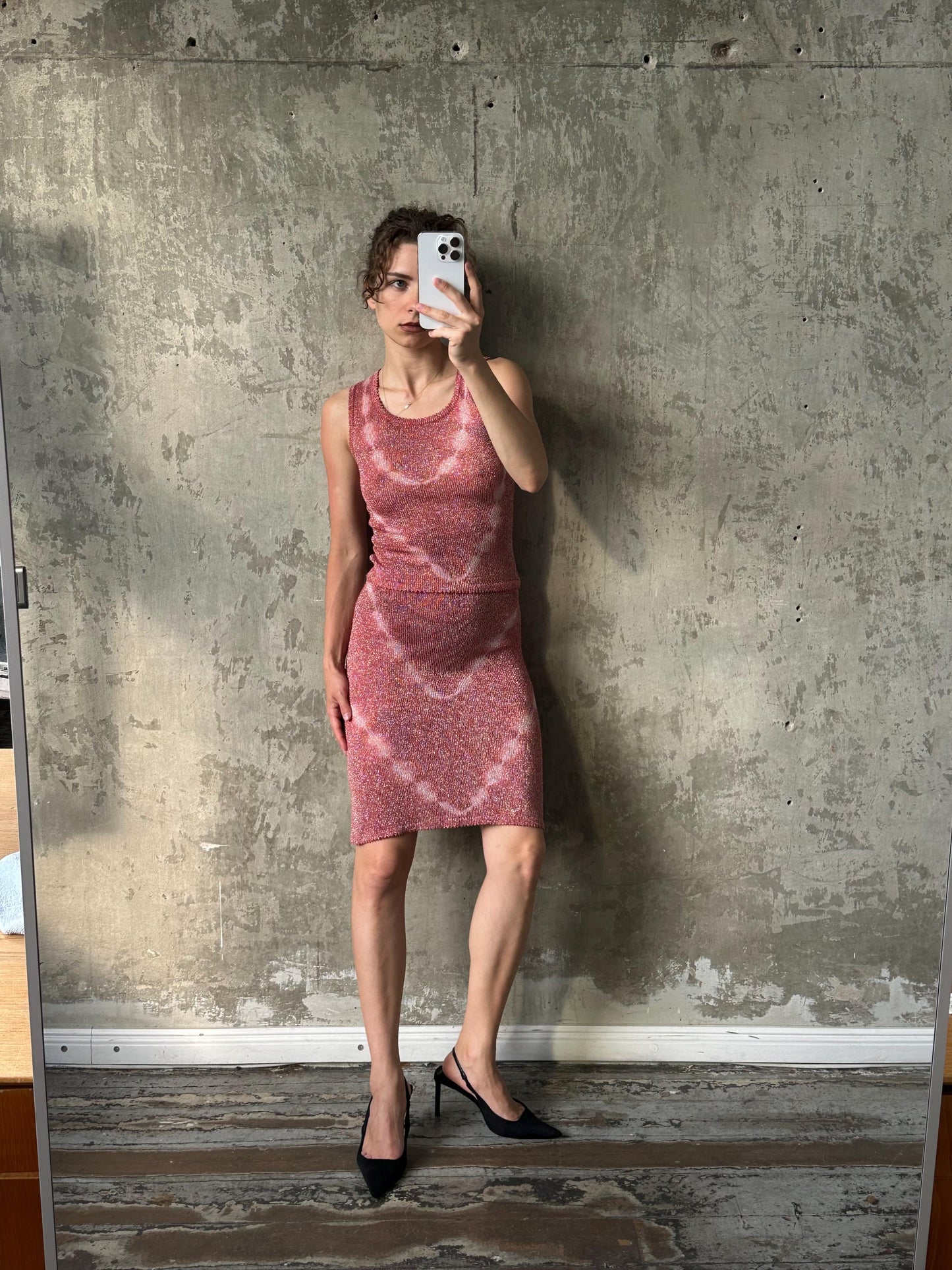 A woman in a pink dress is taking a selfie in front of a concrete wall.