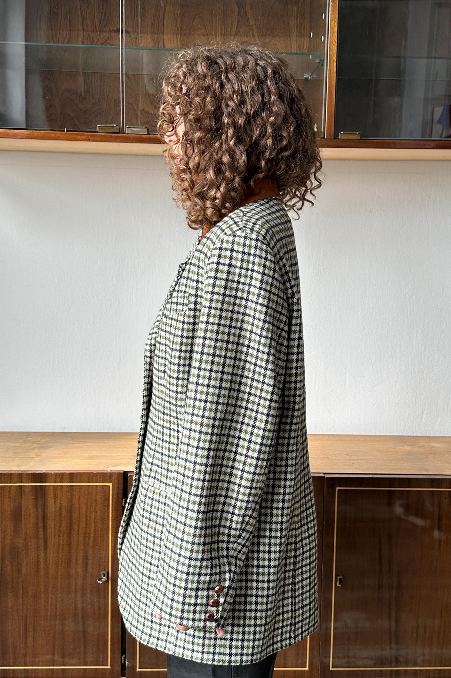 A woman with curly hair wearing a checkered jacket stands in front of a wooden cabinet.
