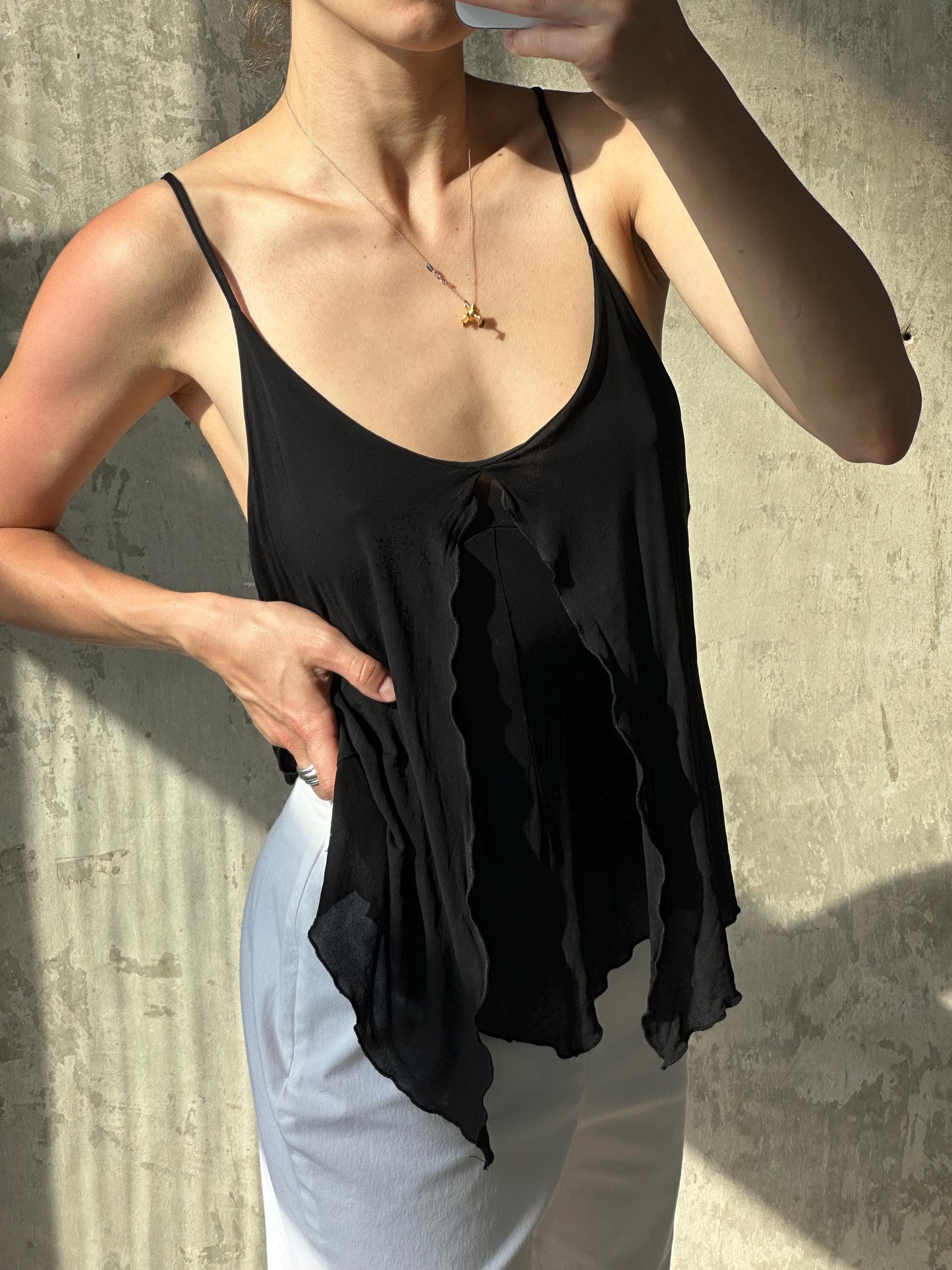 A woman wearing a black top and white pants is taking a selfie with her phone, holding it up to her face.