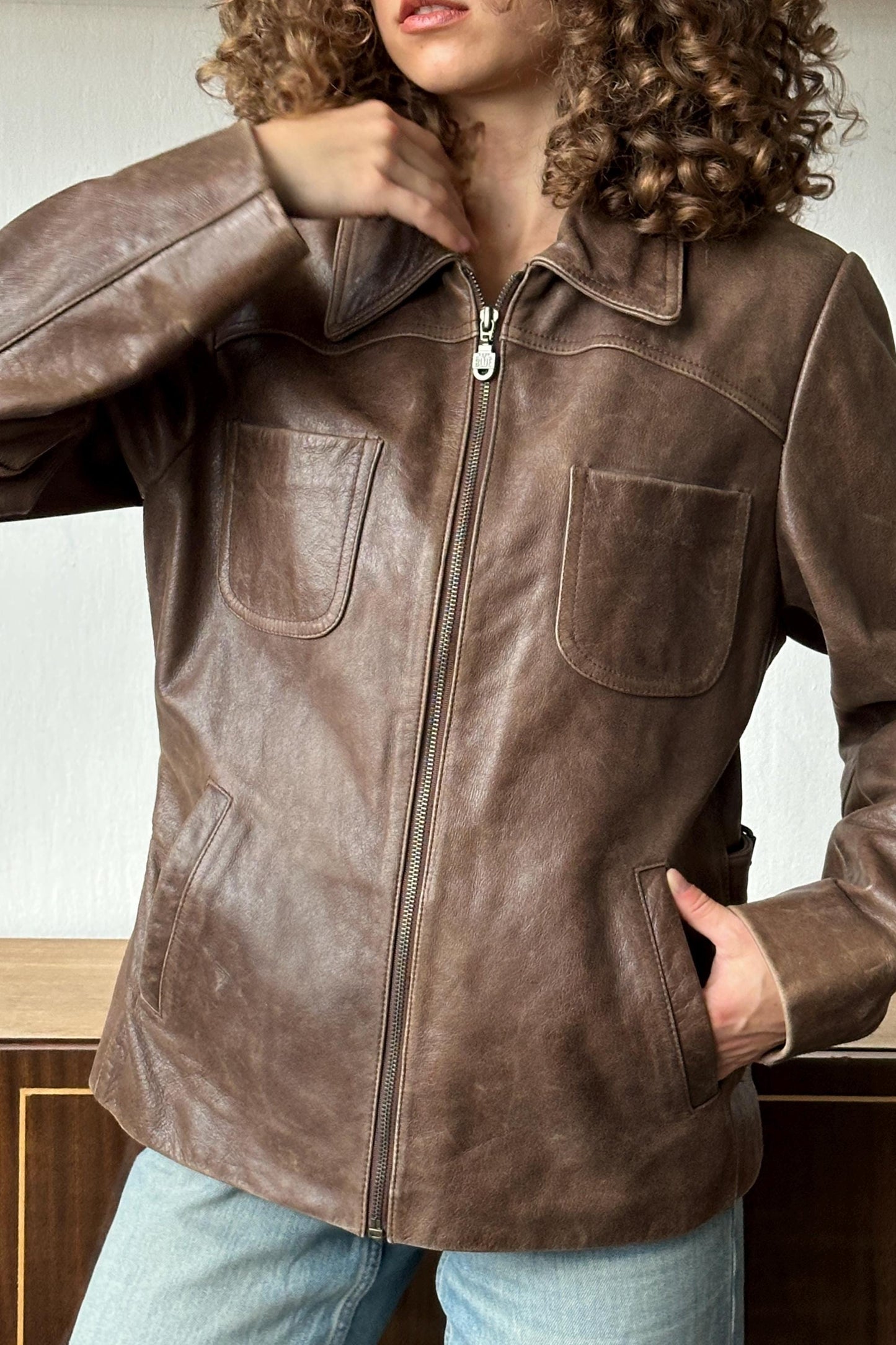 A person wearing a brown leather jacket with a zipper and two pockets, standing in front of a wooden cabinet.