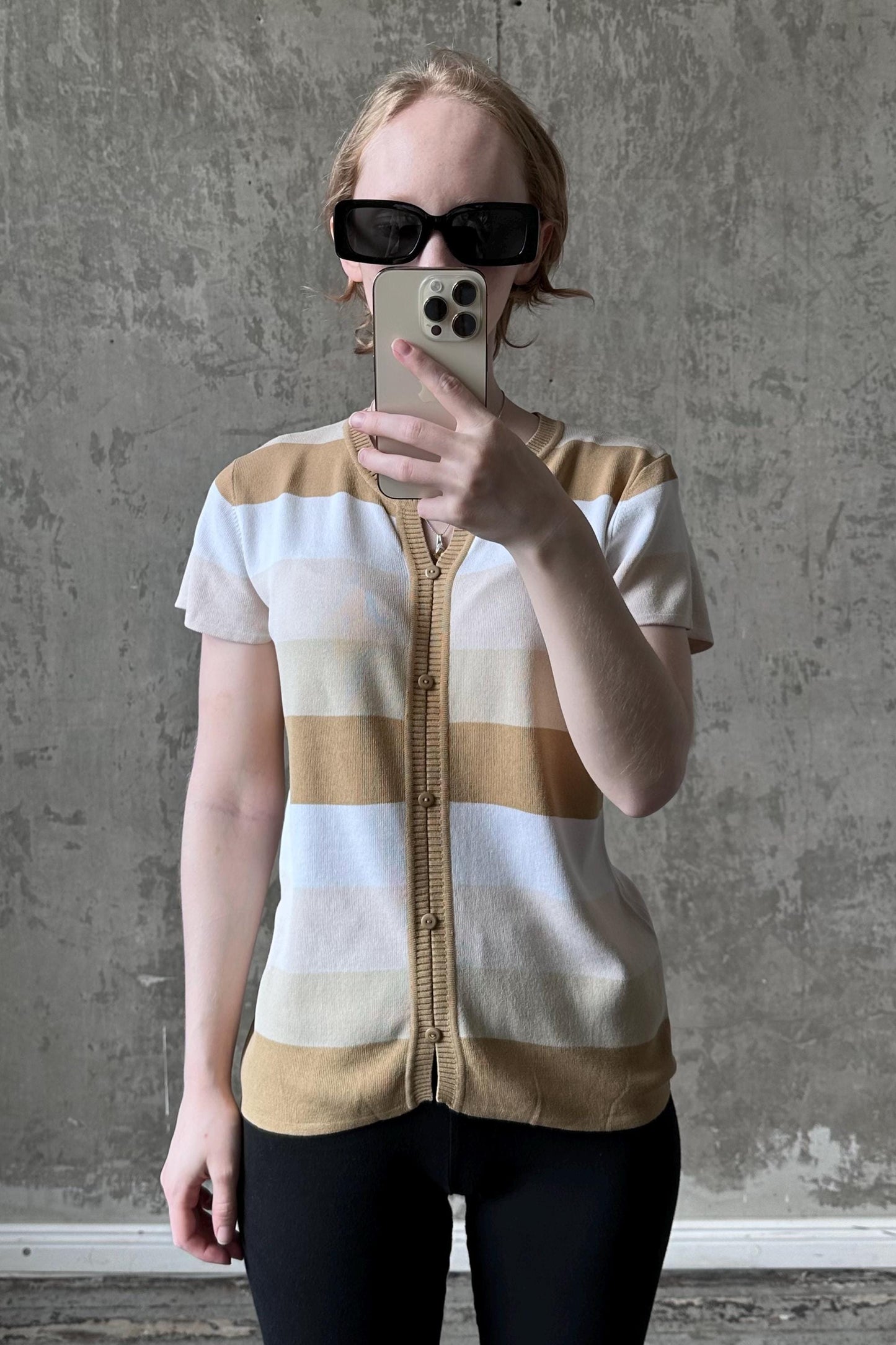 A woman wearing a striped sweater and sunglasses is taking a selfie with her iPhone against a concrete wall.