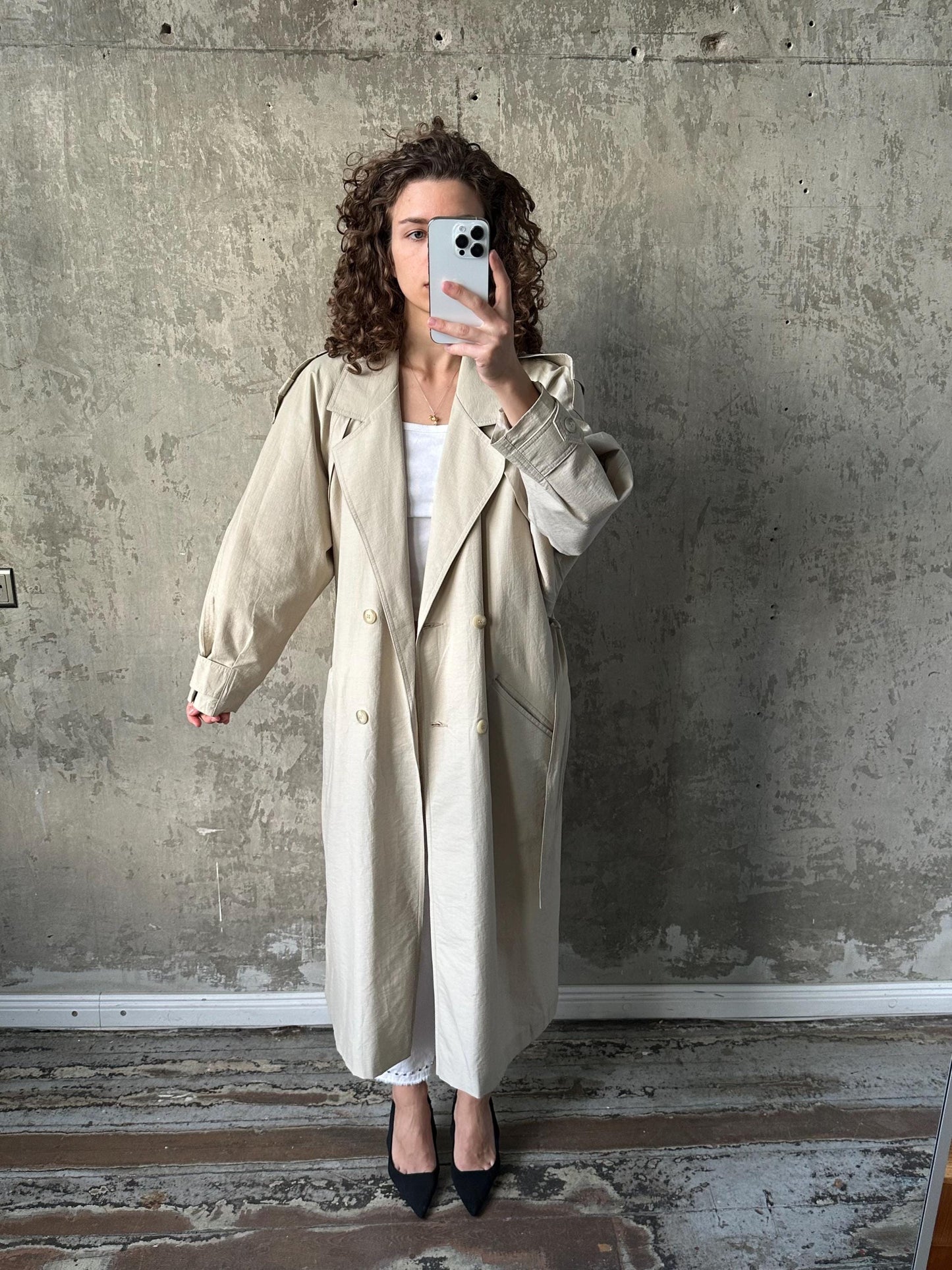 A woman in a beige trench coat is taking a selfie with a smartphone, standing in front of a concrete wall.