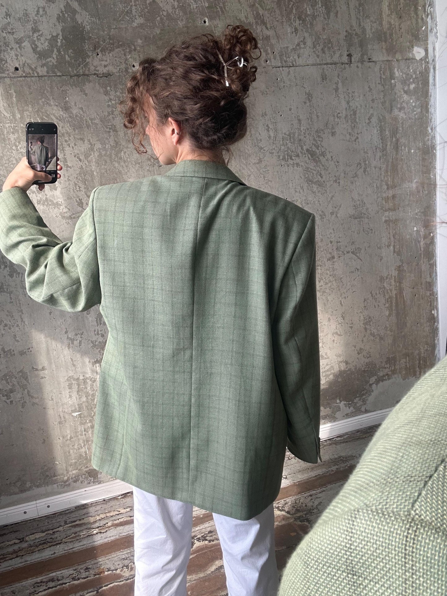 A person is taking a selfie with a smartphone, wearing a green plaid blazer and white pants.