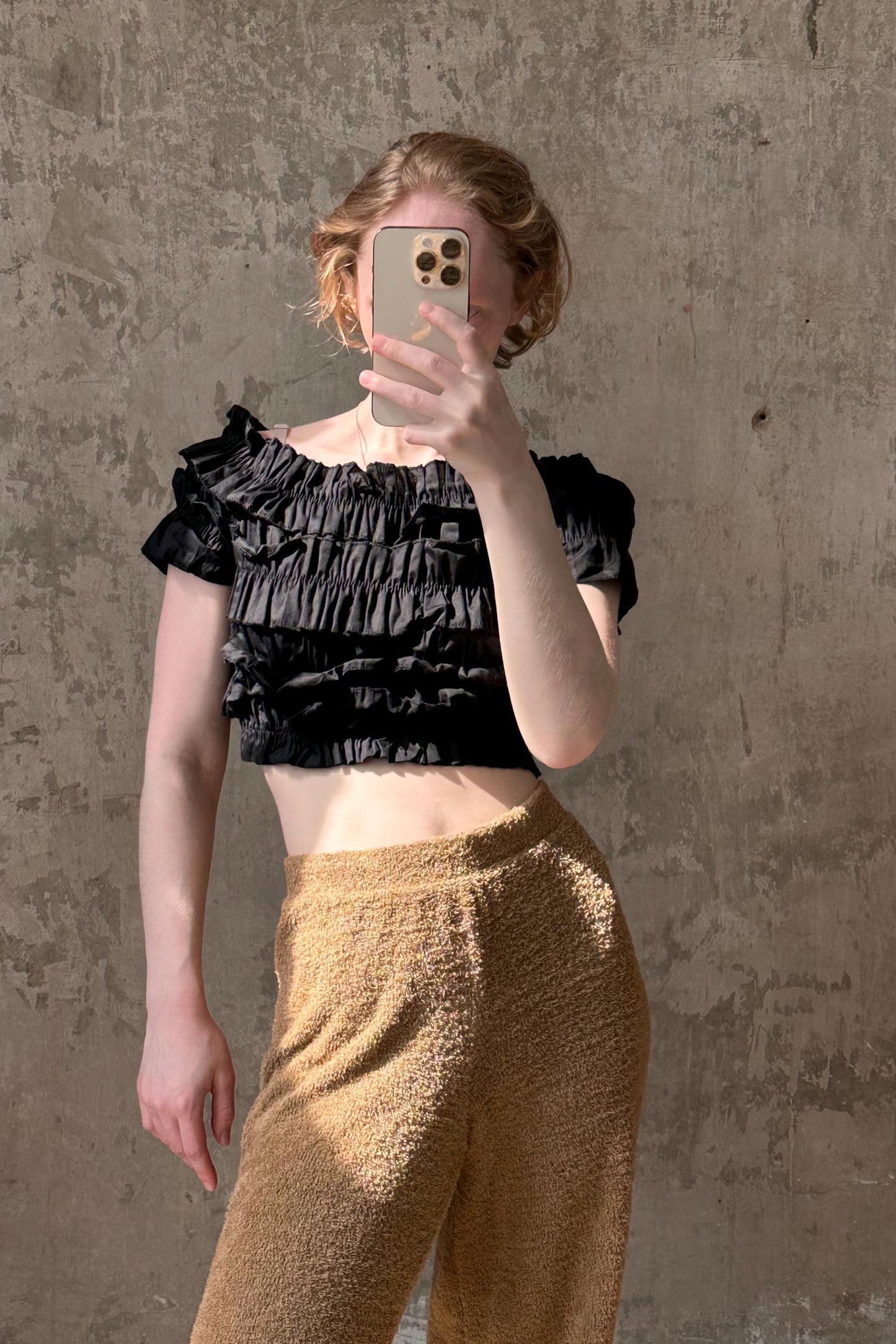 A person is taking a selfie with a smartphone, wearing a black ruffled top and beige pants, against a concrete wall background.