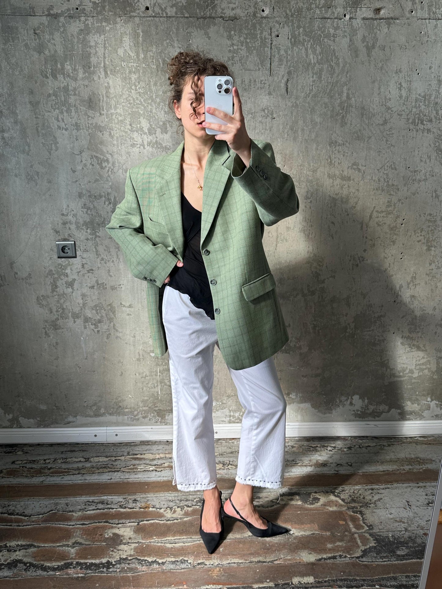 A woman in a green blazer and white pants is taking a selfie with a white phone, standing in front of a concrete wall.