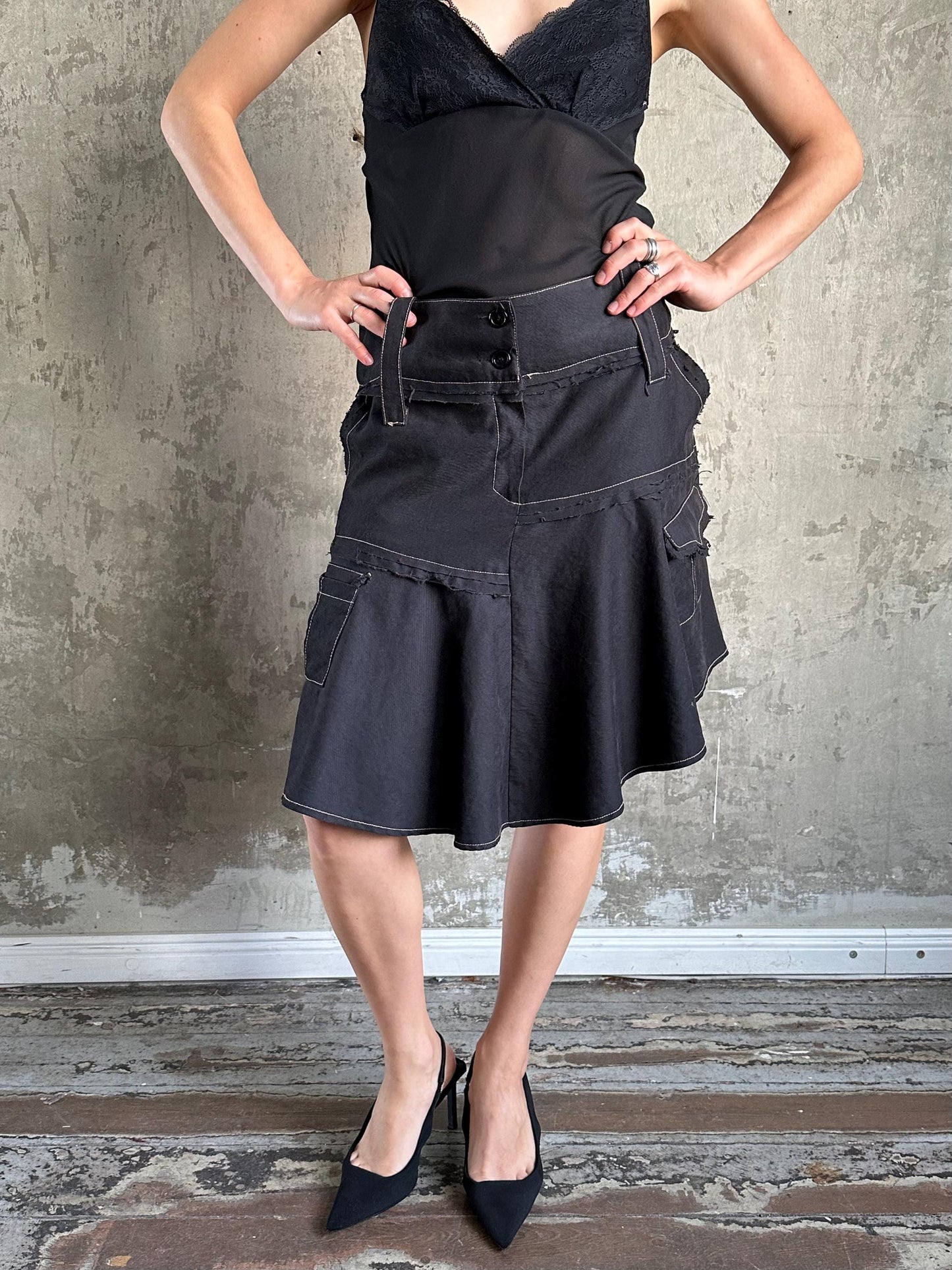 A woman wearing a black skirt and top stands confidently with her hands on her hips against a concrete wall.