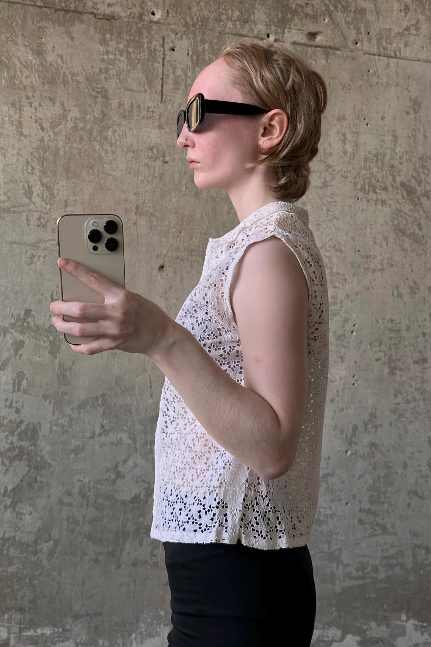 A woman wearing a white lace top and sunglasses is holding a smartphone and looking at the screen.