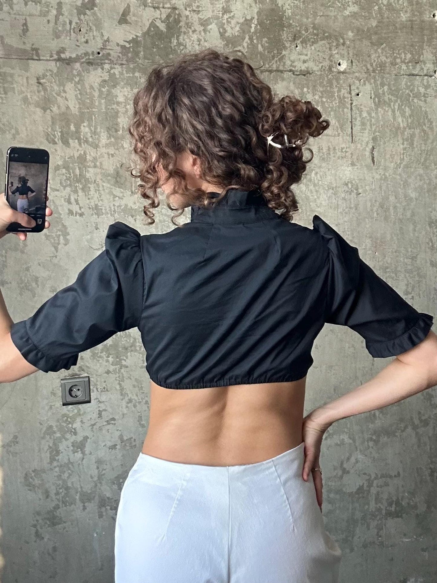 a person taking a selfie with a smartphone, wearing a black crop top and white pants.