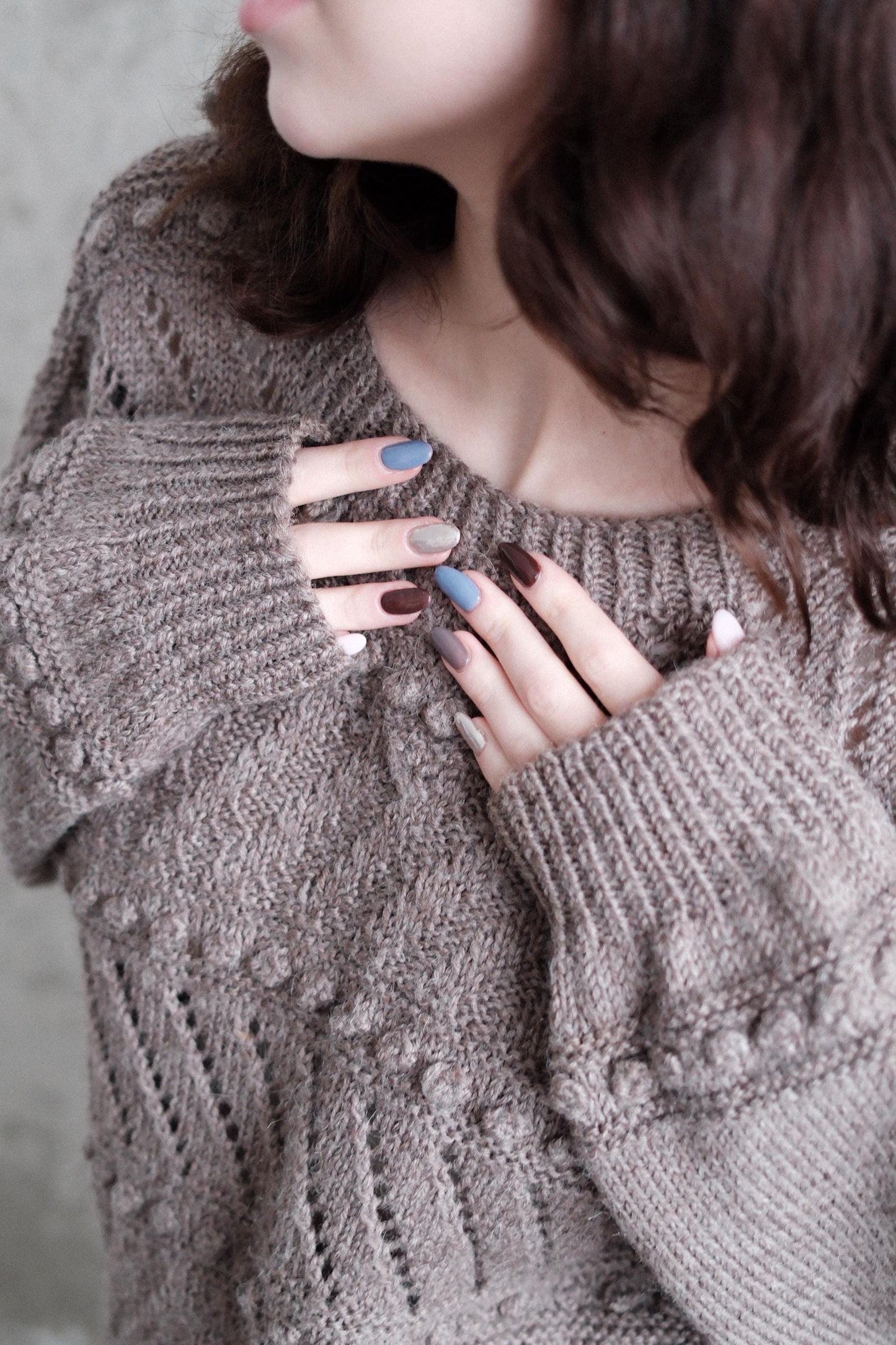 A close-up view of a person's hands and arms wearing a gray knitted sweater, with their nails painted in various colors.