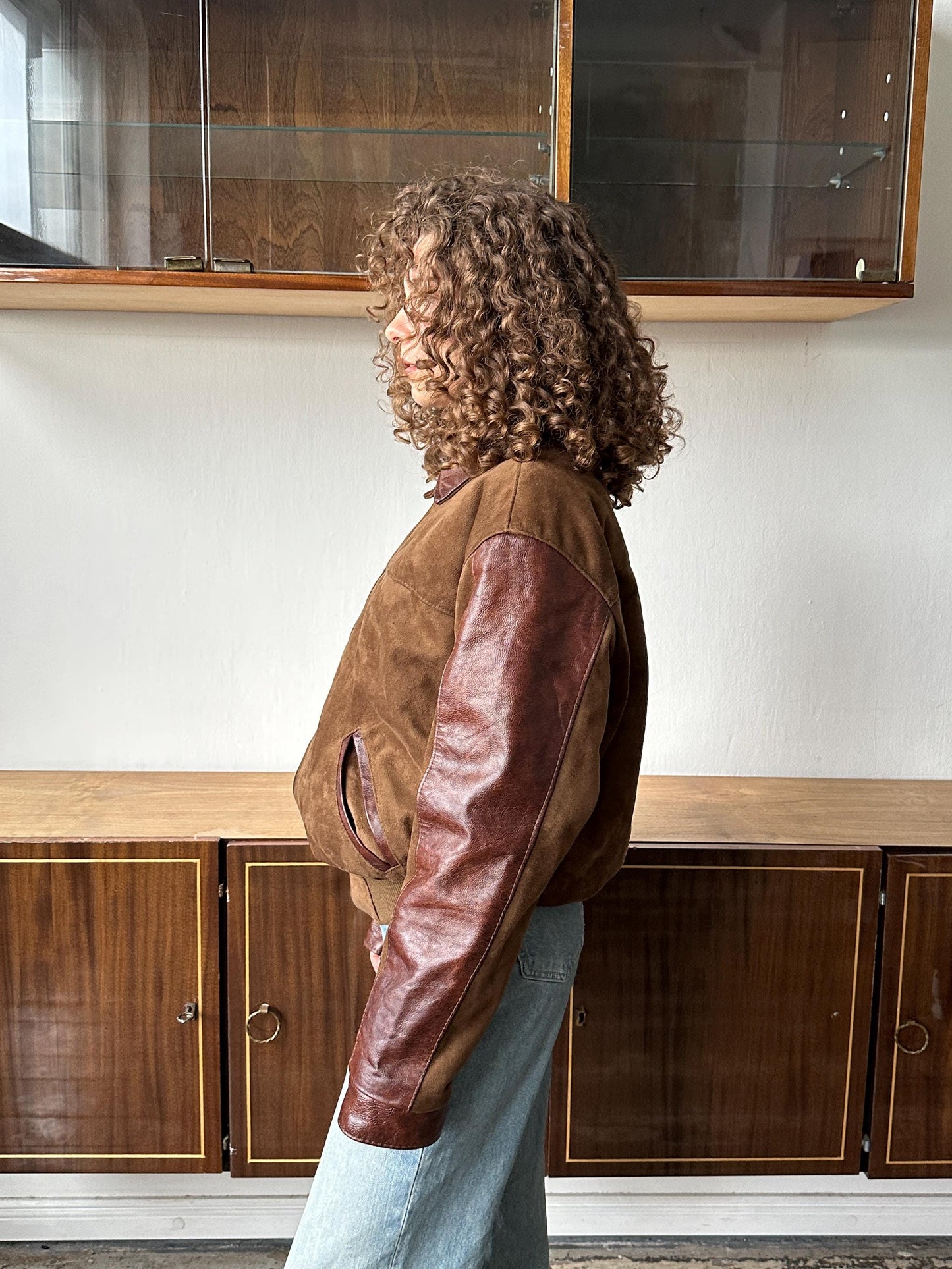 A person with curly hair wearing a brown leather jacket stands in a room with wooden cabinets.