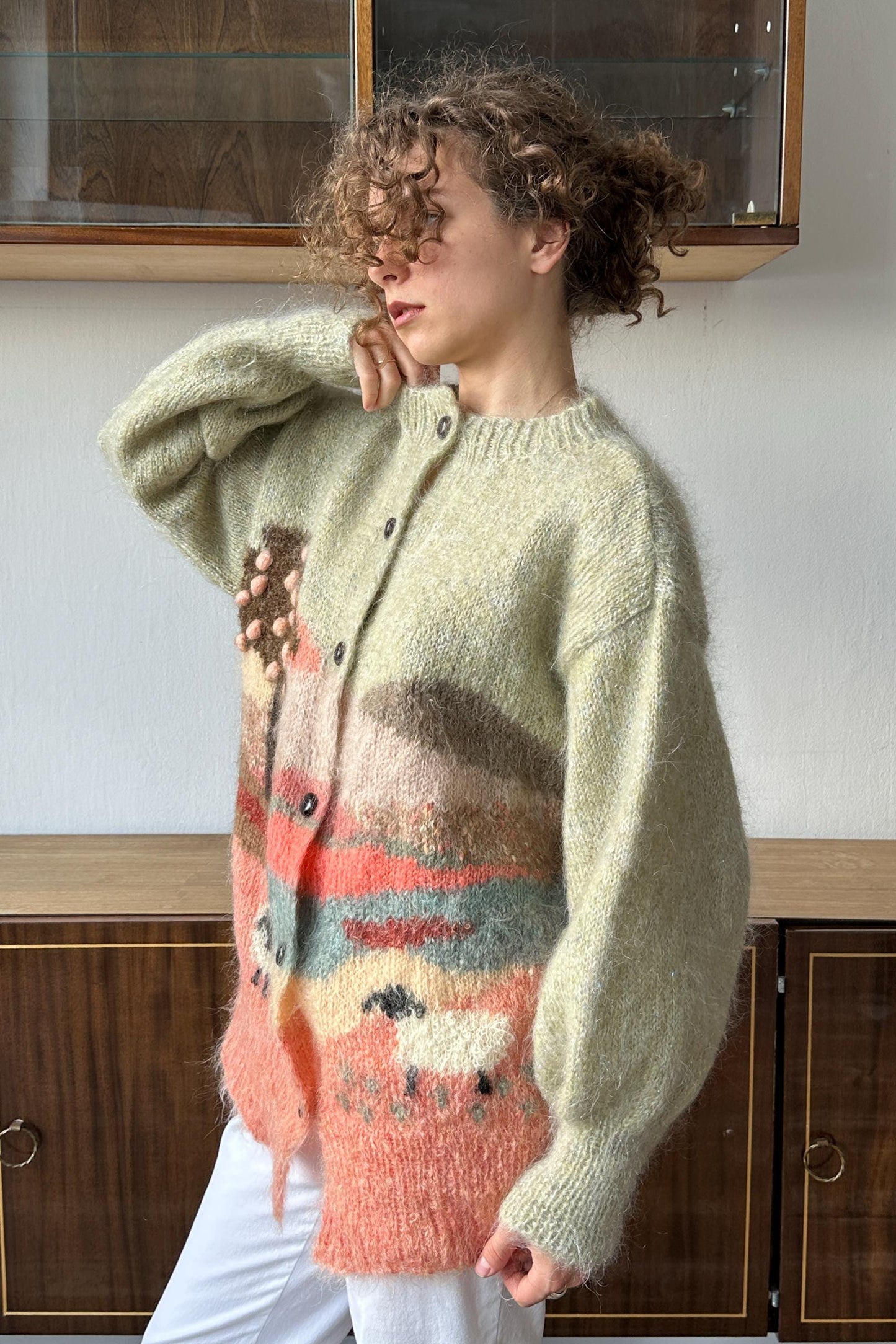 A woman wearing a light pink and orange knitted cardigan stands in front of a wooden cabinet.