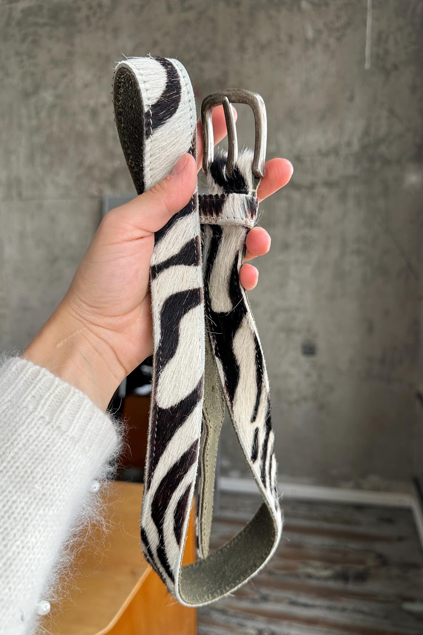 A hand holding a black and white zebra-striped leather belt against a concrete wall background.