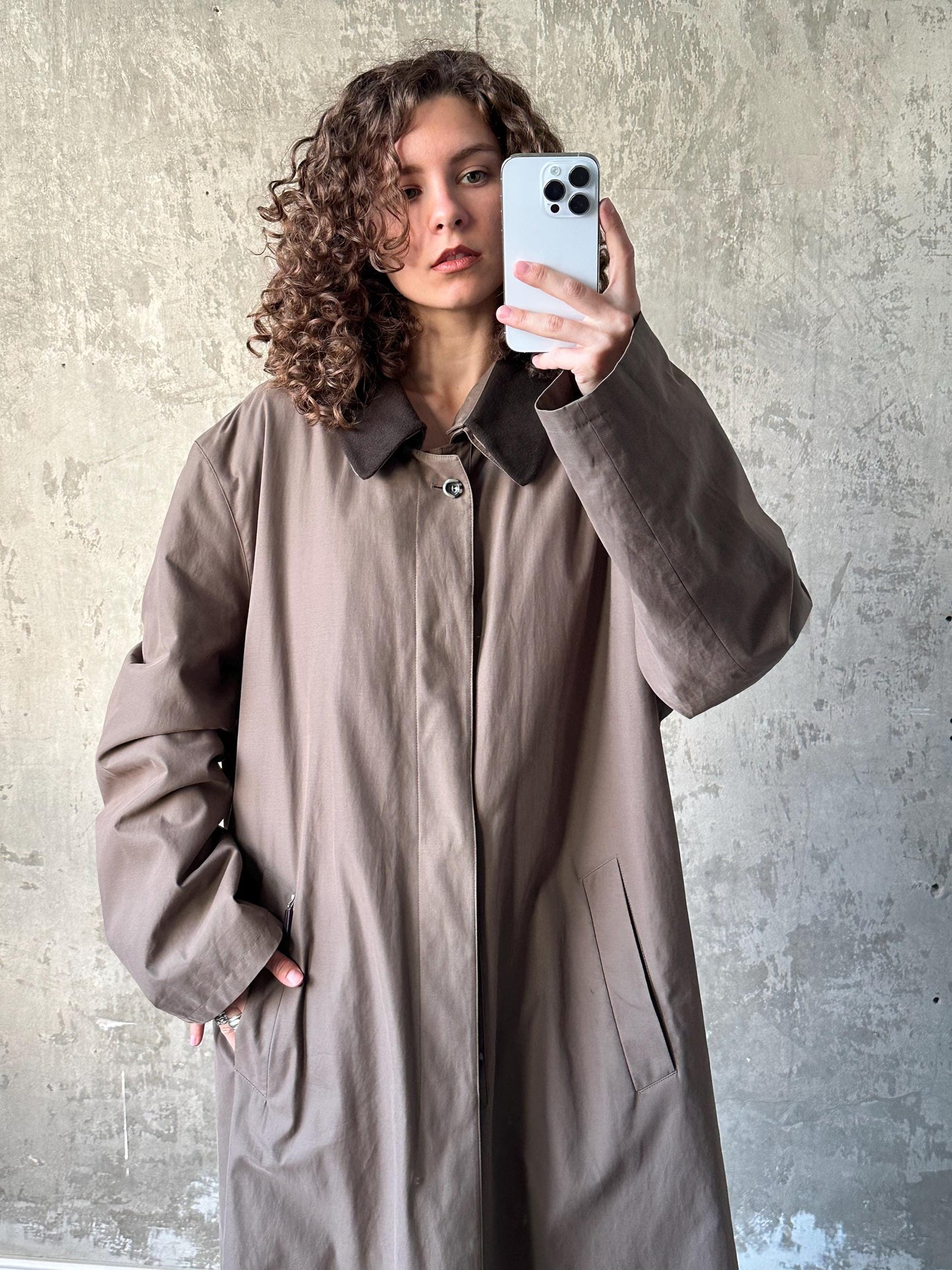 A woman with curly hair is taking a selfie with a white smartphone, wearing a beige coat.