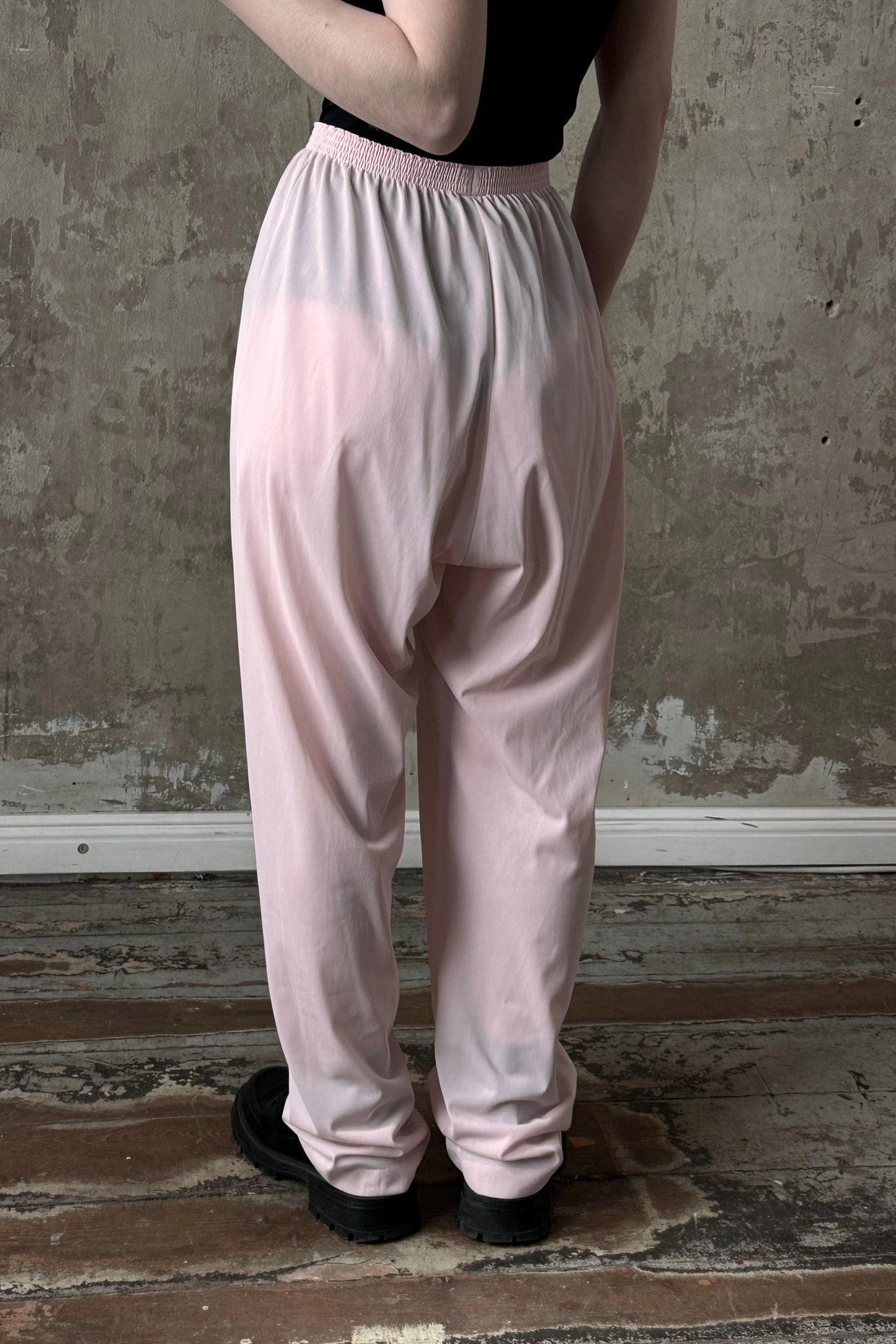 A person wearing a black top and pink pants stands in front of a concrete wall.
