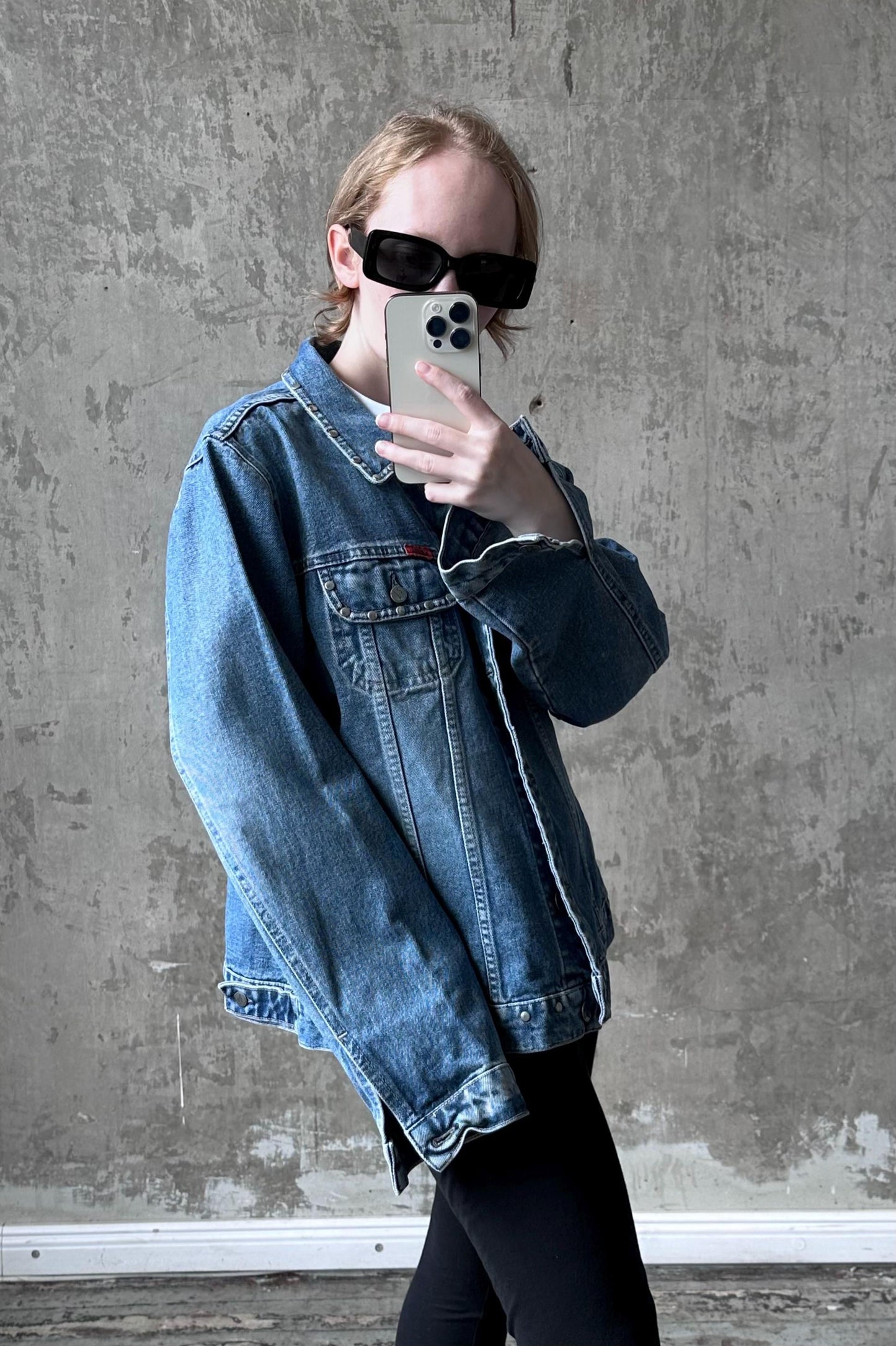 A person wearing a denim jacket and sunglasses is taking a selfie with a smartphone.