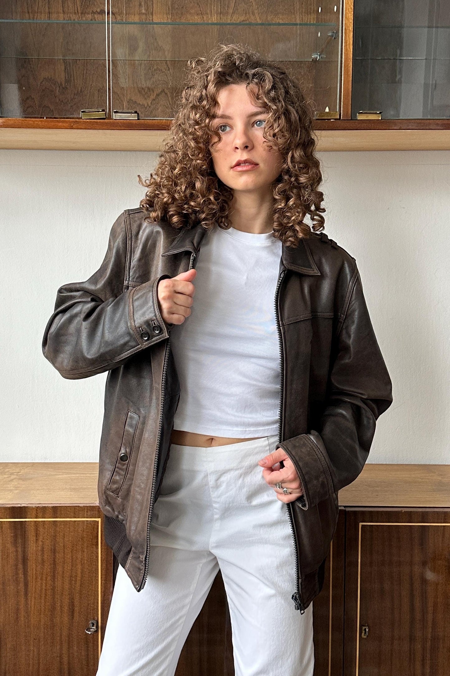 A woman with curly hair wearing a brown leather jacket and white pants stands in front of a wooden cabinet.