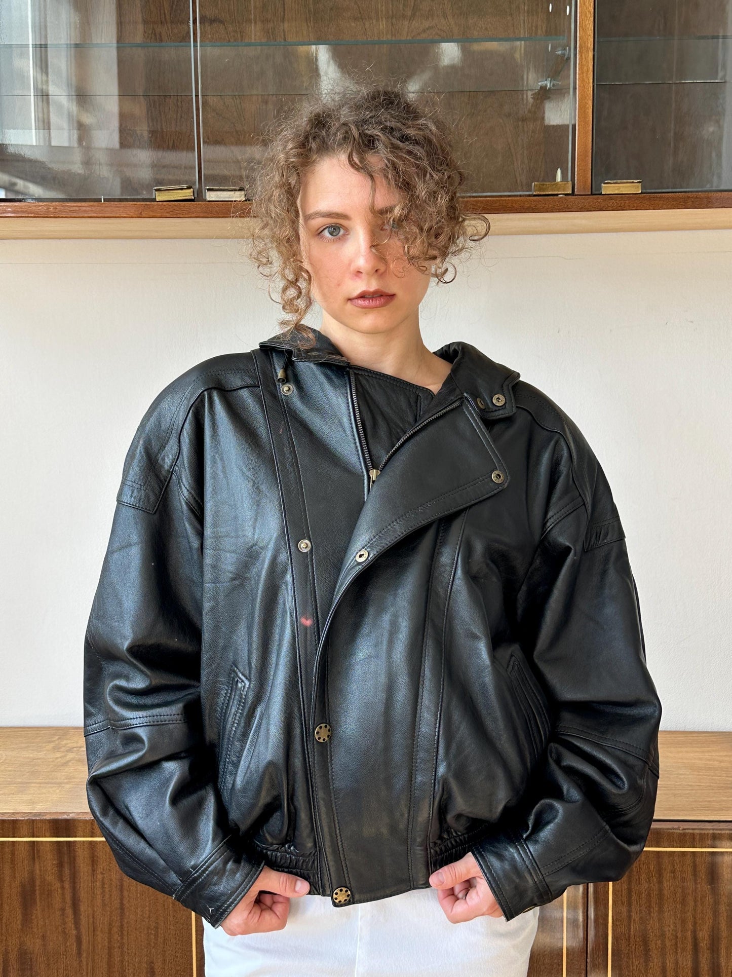 A woman with curly hair wearing a black leather jacket stands in front of a glass display case.