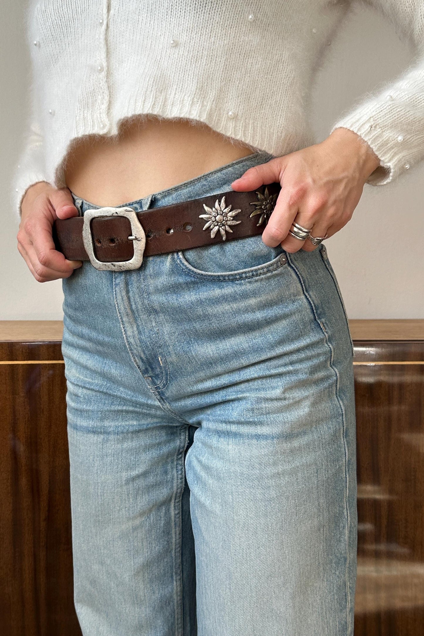 A person wearing a white sweater and blue jeans, with a brown belt featuring silver stars, stands in front of a wooden cabinet.