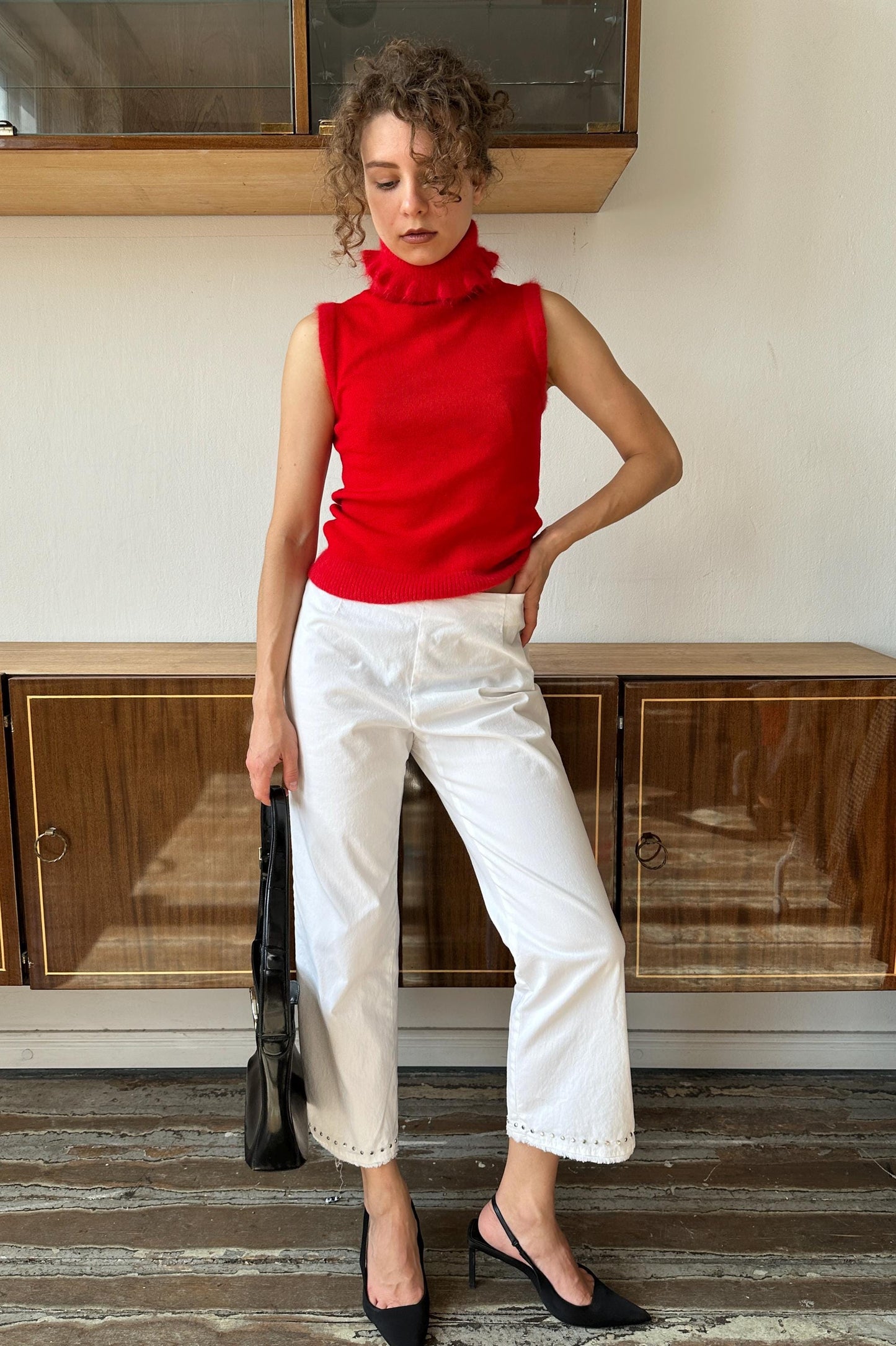 A woman wearing a red sleeveless top and white pants stands in a room with wooden cabinets in the background.