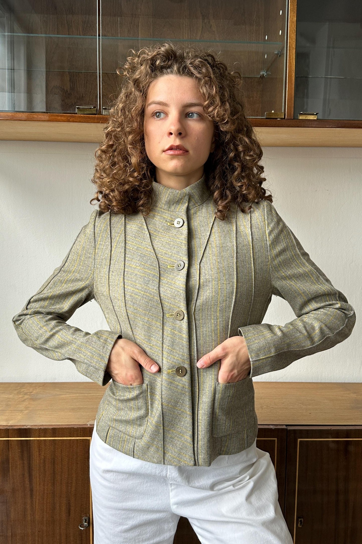 A woman with curly hair wearing a beige jacket and white pants stands in front of a wooden cabinet.