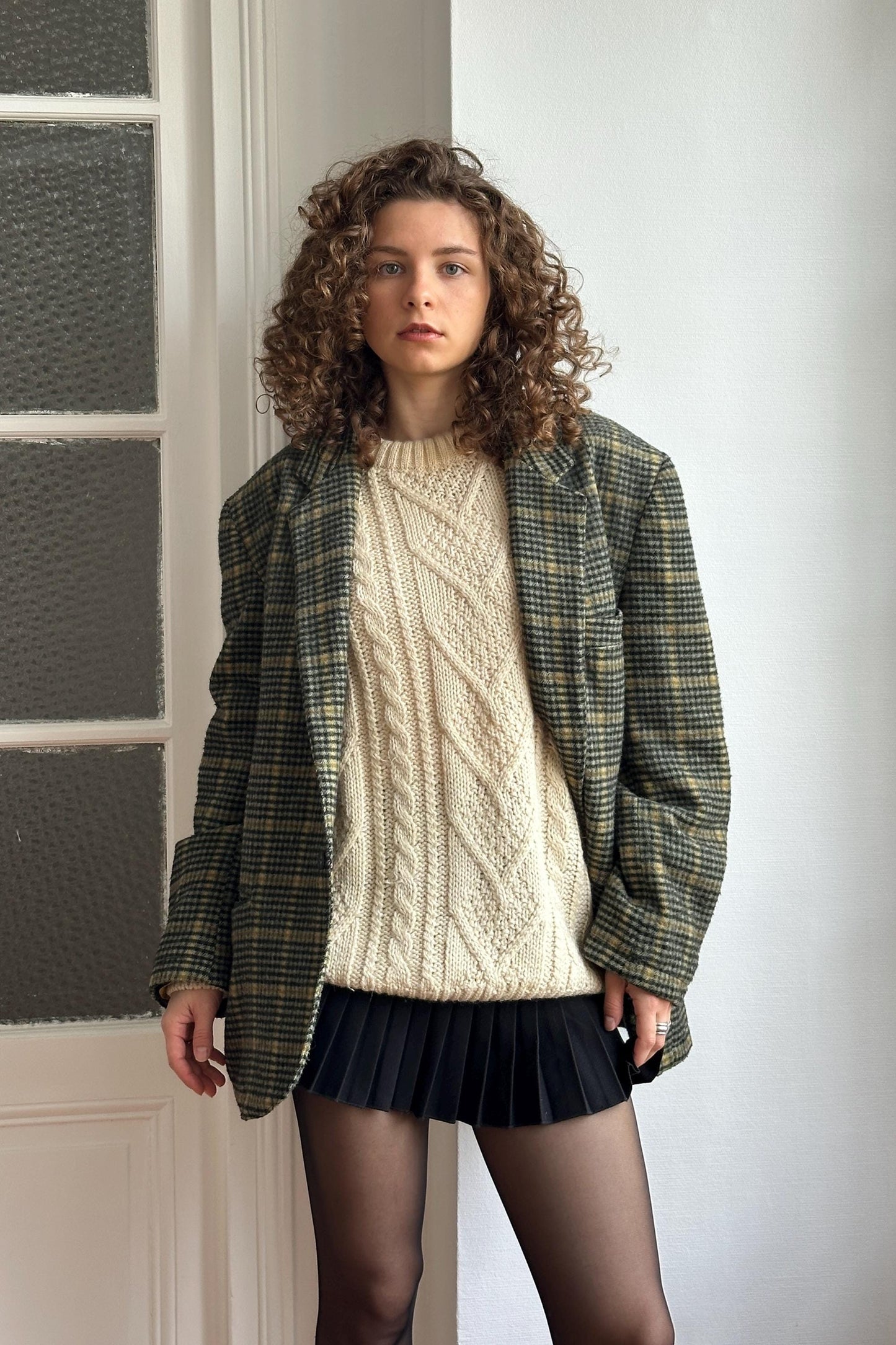A young woman with curly hair stands in front of a white door, wearing a plaid jacket, a white sweater, and a black skirt.