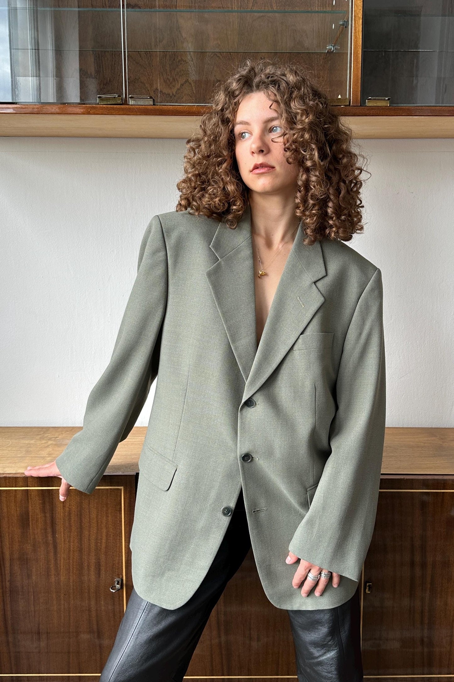 A woman with curly hair wearing a gray blazer stands in front of a wooden cabinet.