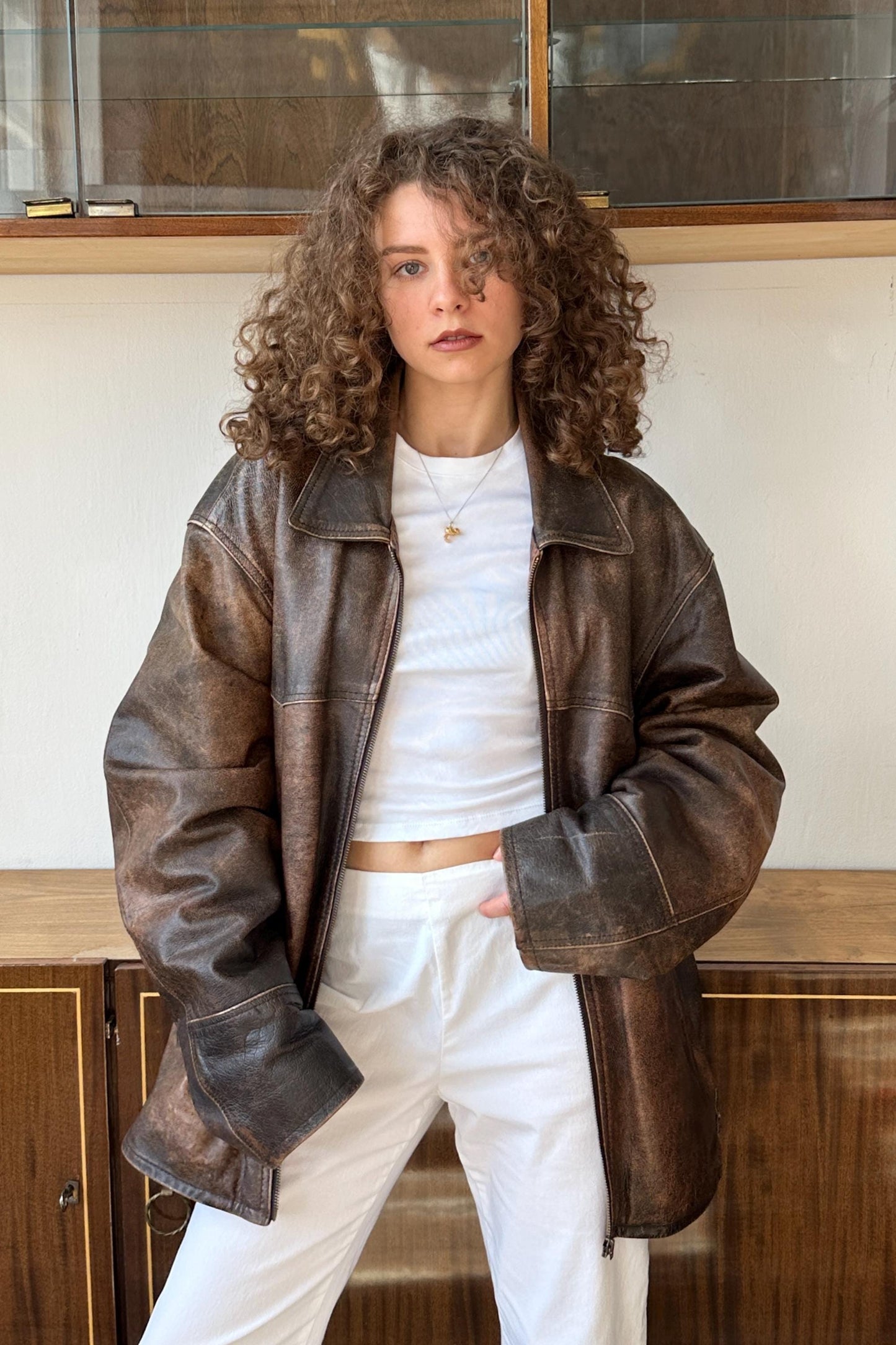 A young woman with curly hair wearing a brown leather jacket and white pants stands in front of a glass display case.