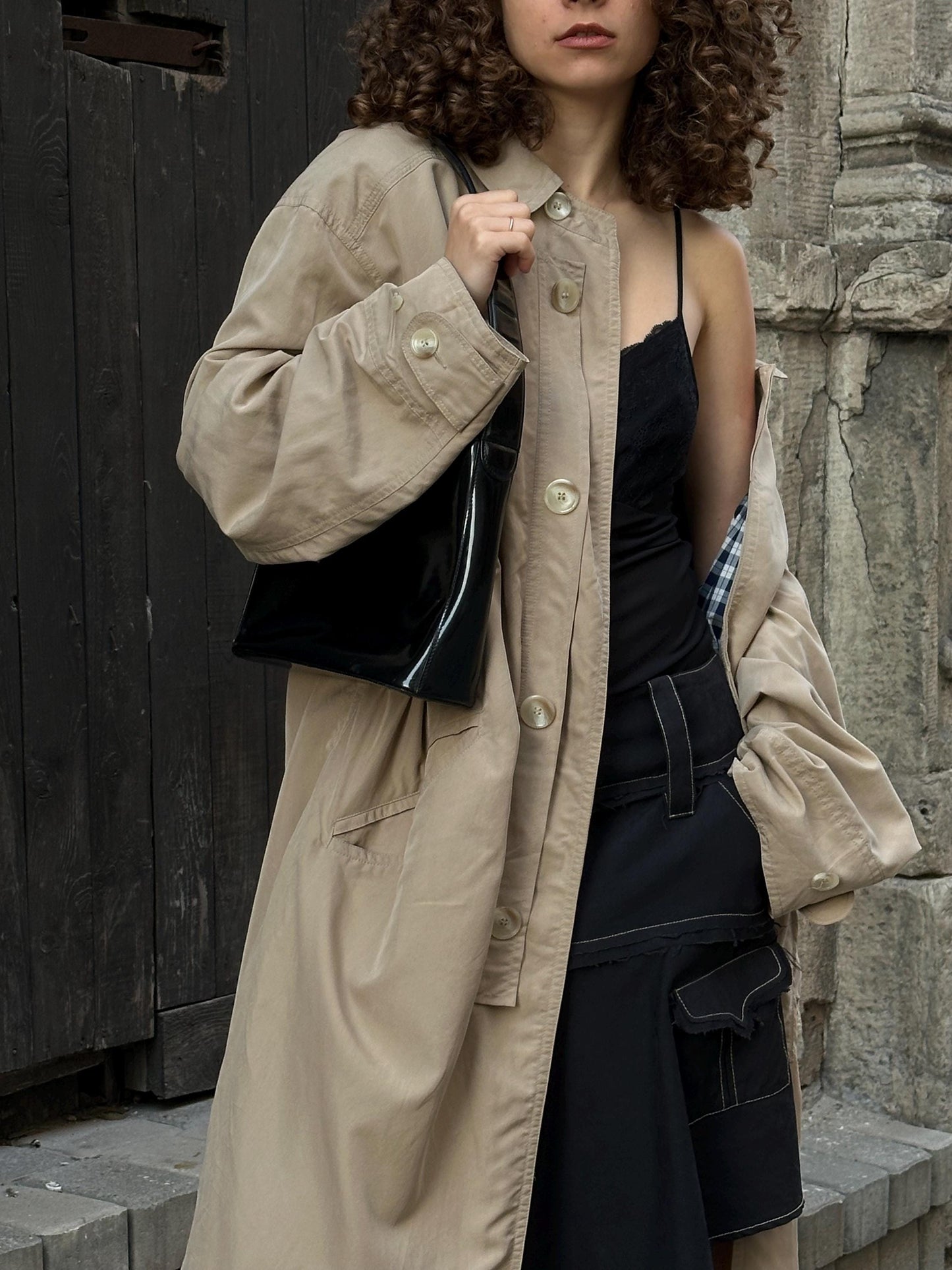 A woman wearing a beige trench coat and black skirt stands in front of a stone wall, holding a black purse.